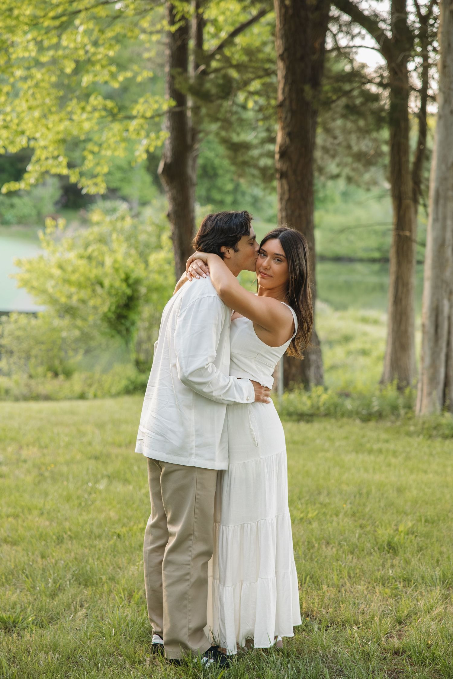 Couple embracing in a lush green park near trees and a lake at golden hour.