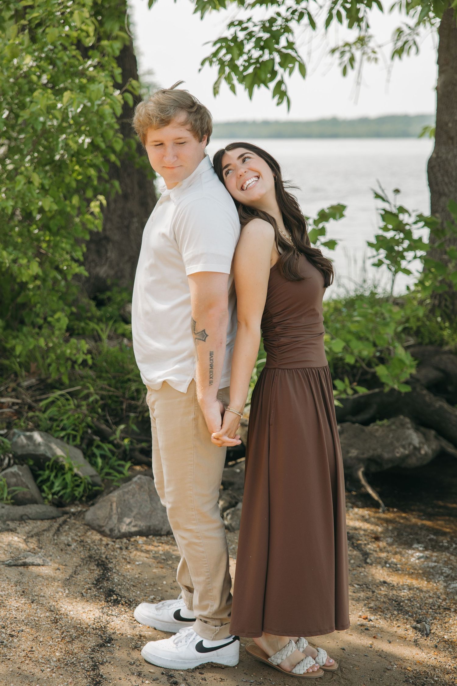 Couple holding hands back-to-back near a lakeside, woman in brown maxi dress smiling and looking up.