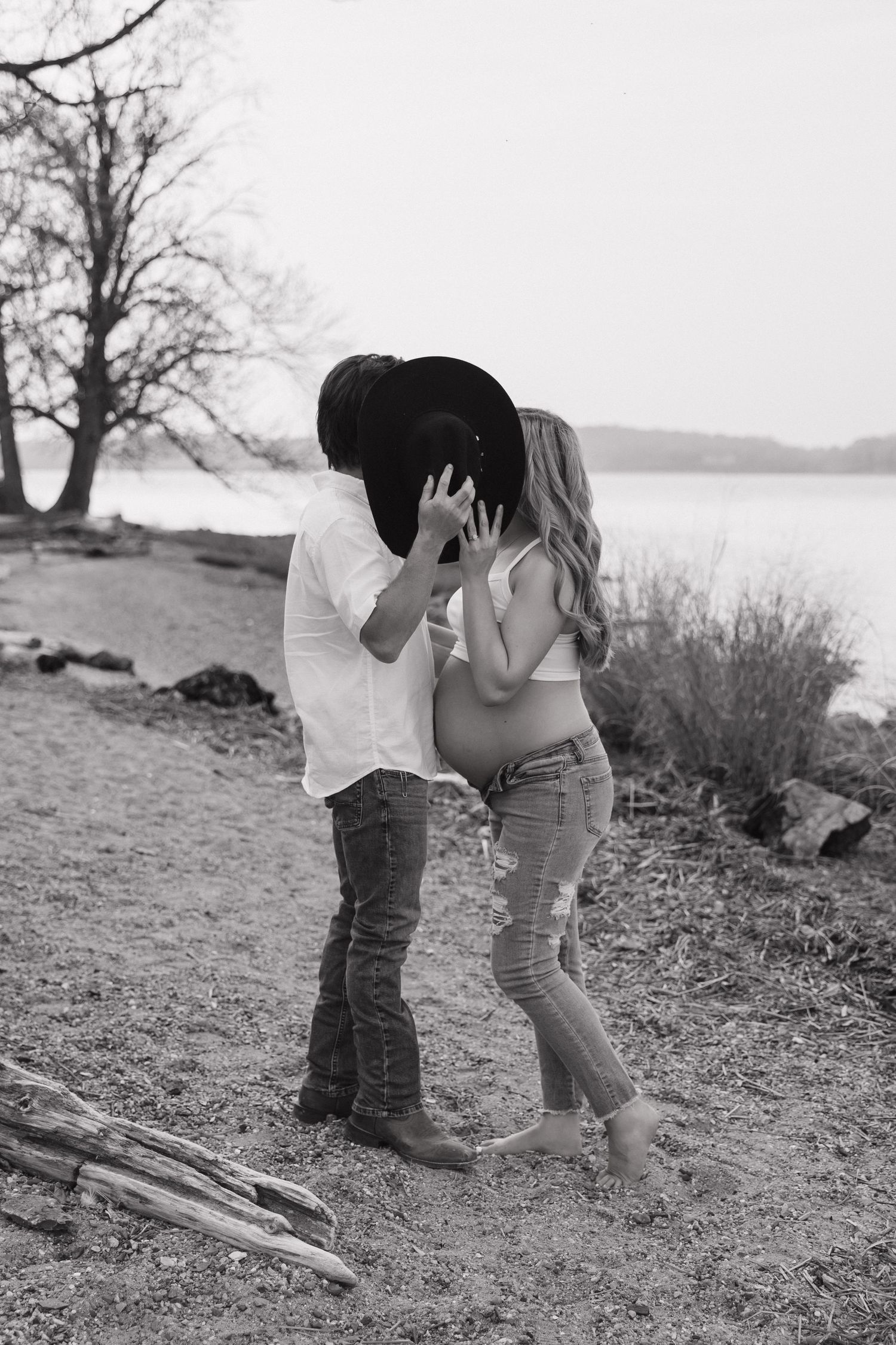 Black and white photo of a couple kissing on a rocky beach, woman showing pregnant belly, man wearing cowboy hat.
