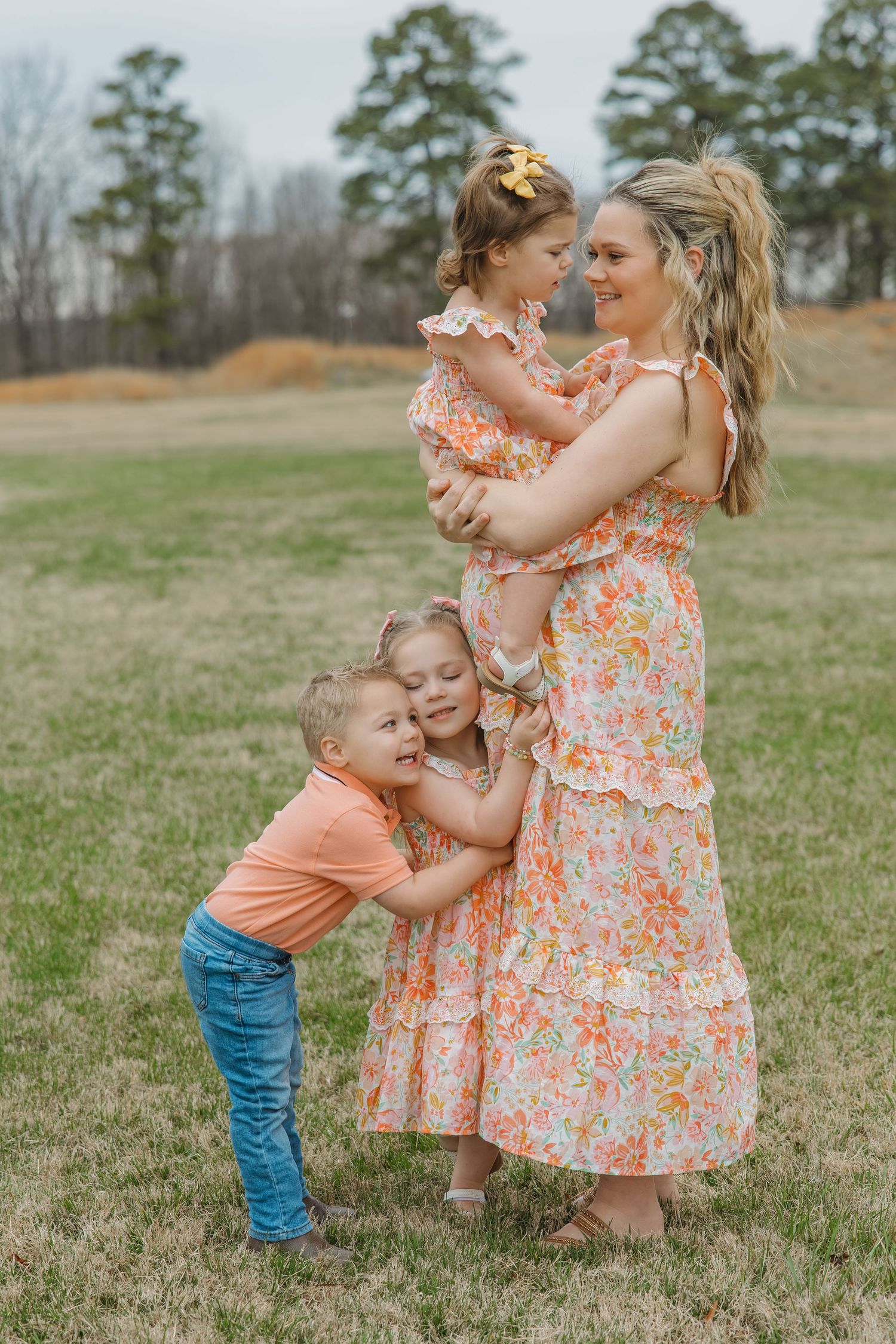 Smiling pregnant mother holds toddler girl while two other young children hug her in a grassy outdoor setting.