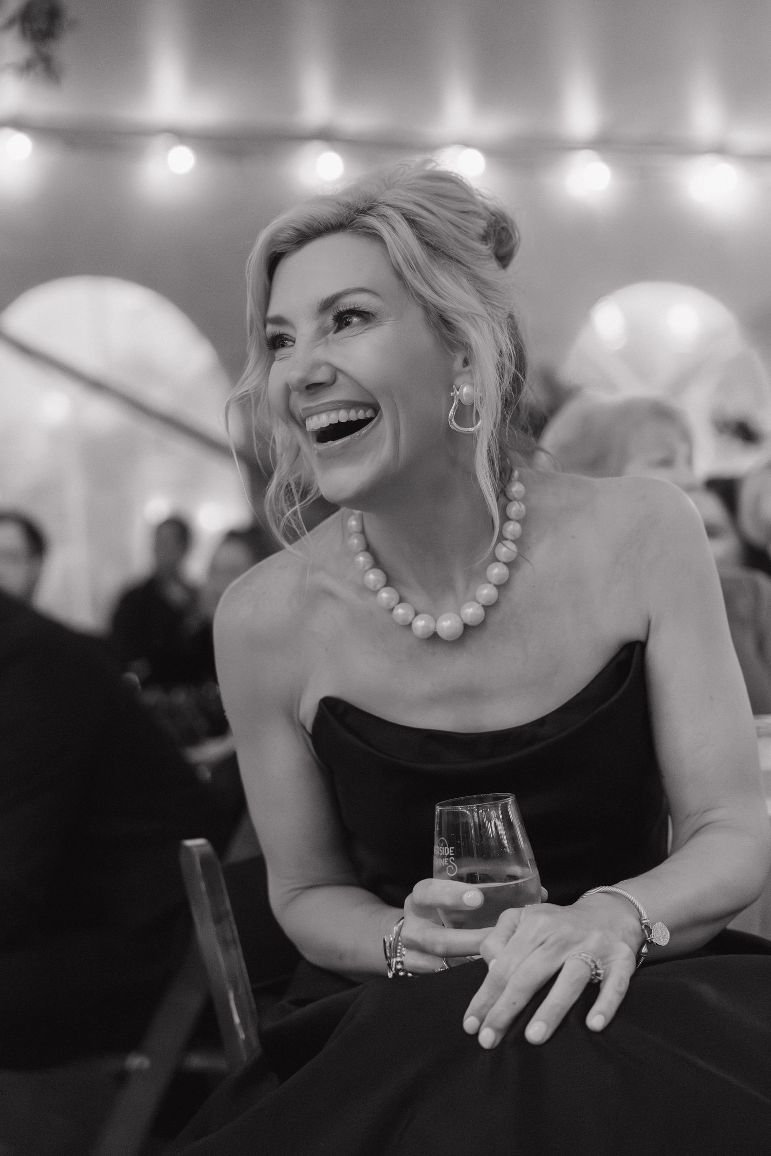 Elegant blonde wedding guest laughs joyfully while holding a drink at a tented wedding reception.