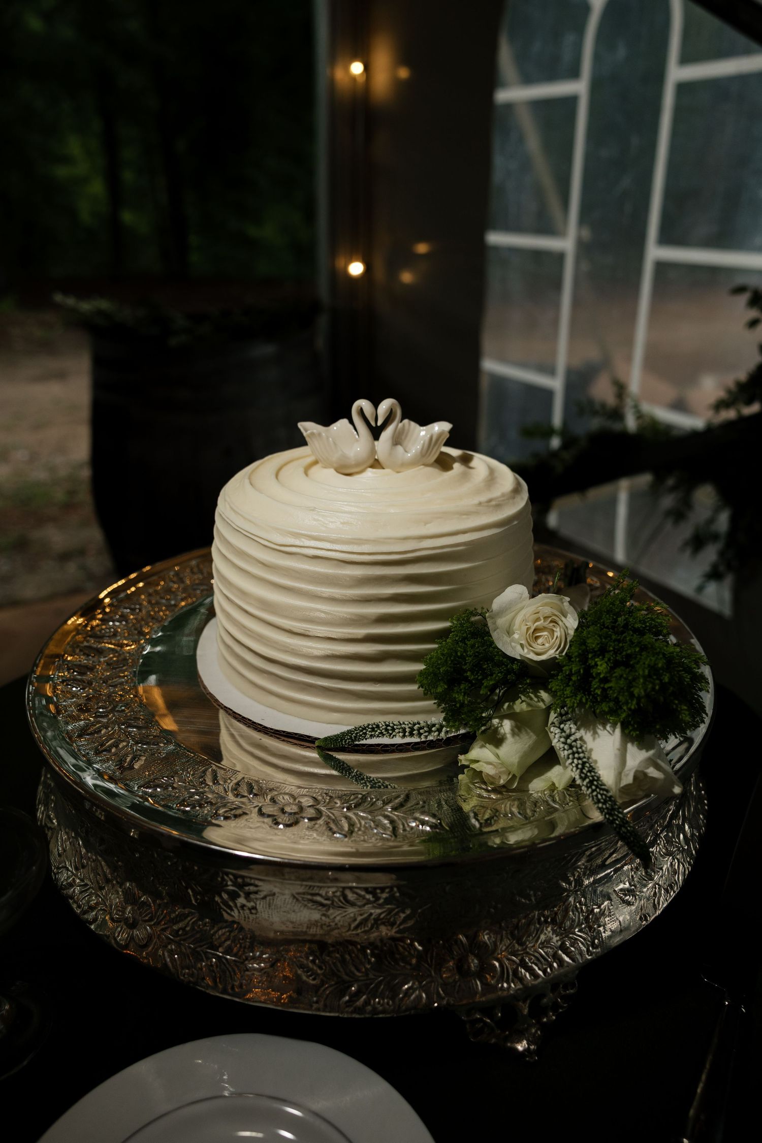 Single-tier white textured wedding cake with swan toppers on silver stand with white rose accents.