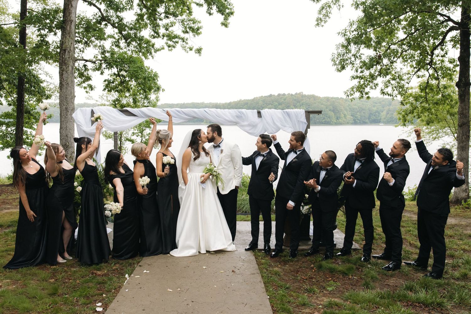 Wedding party cheers as bride and groom kiss outdoors near a lake with trees in background.