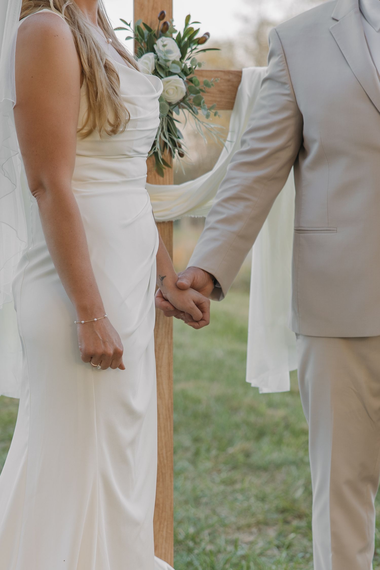 Close-up of bride and groom holding hands at wooden cross altar adorned with white floral arrangement.