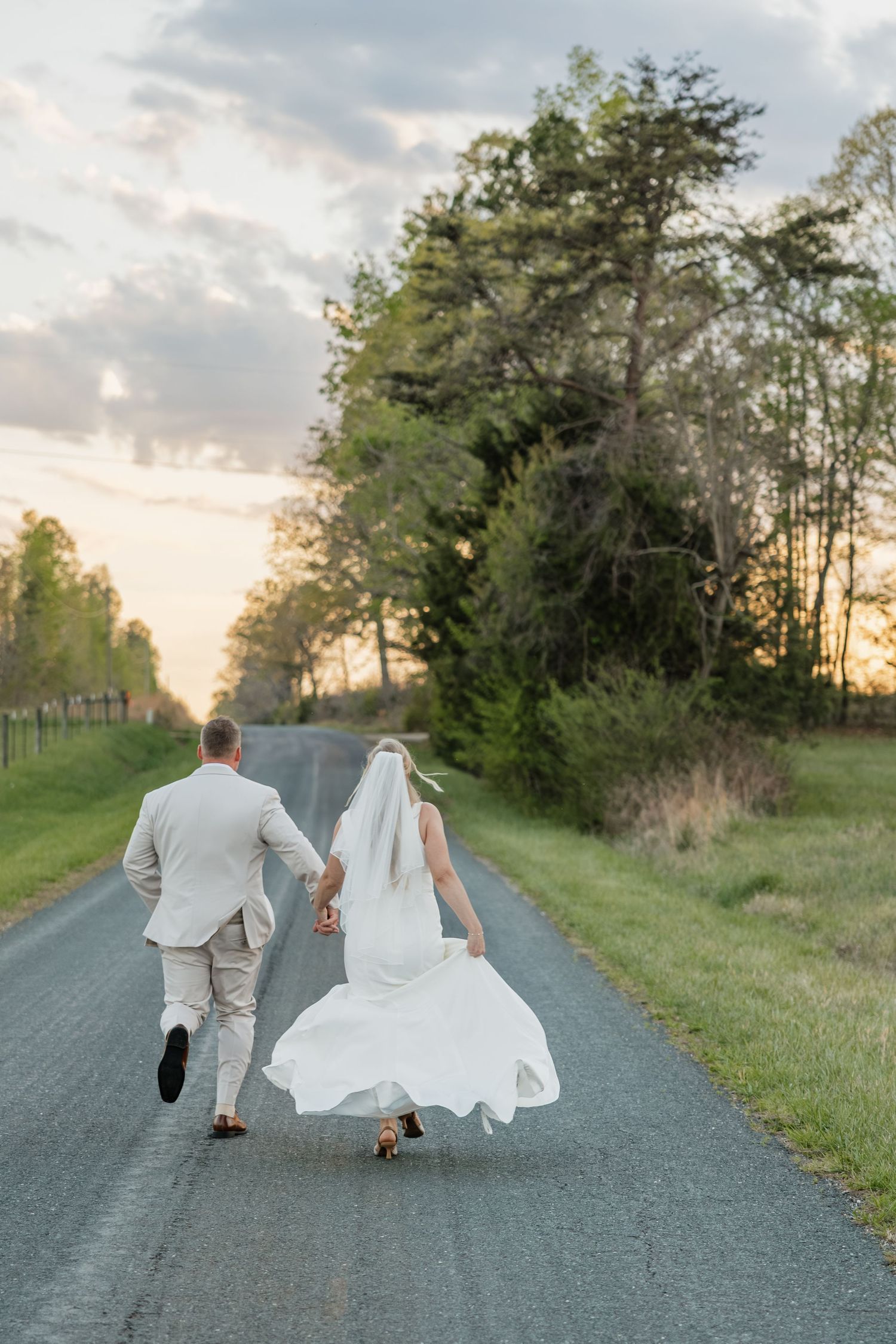 Bride and groom holding hands running down a scenic country road at sunset in wedding attire.
