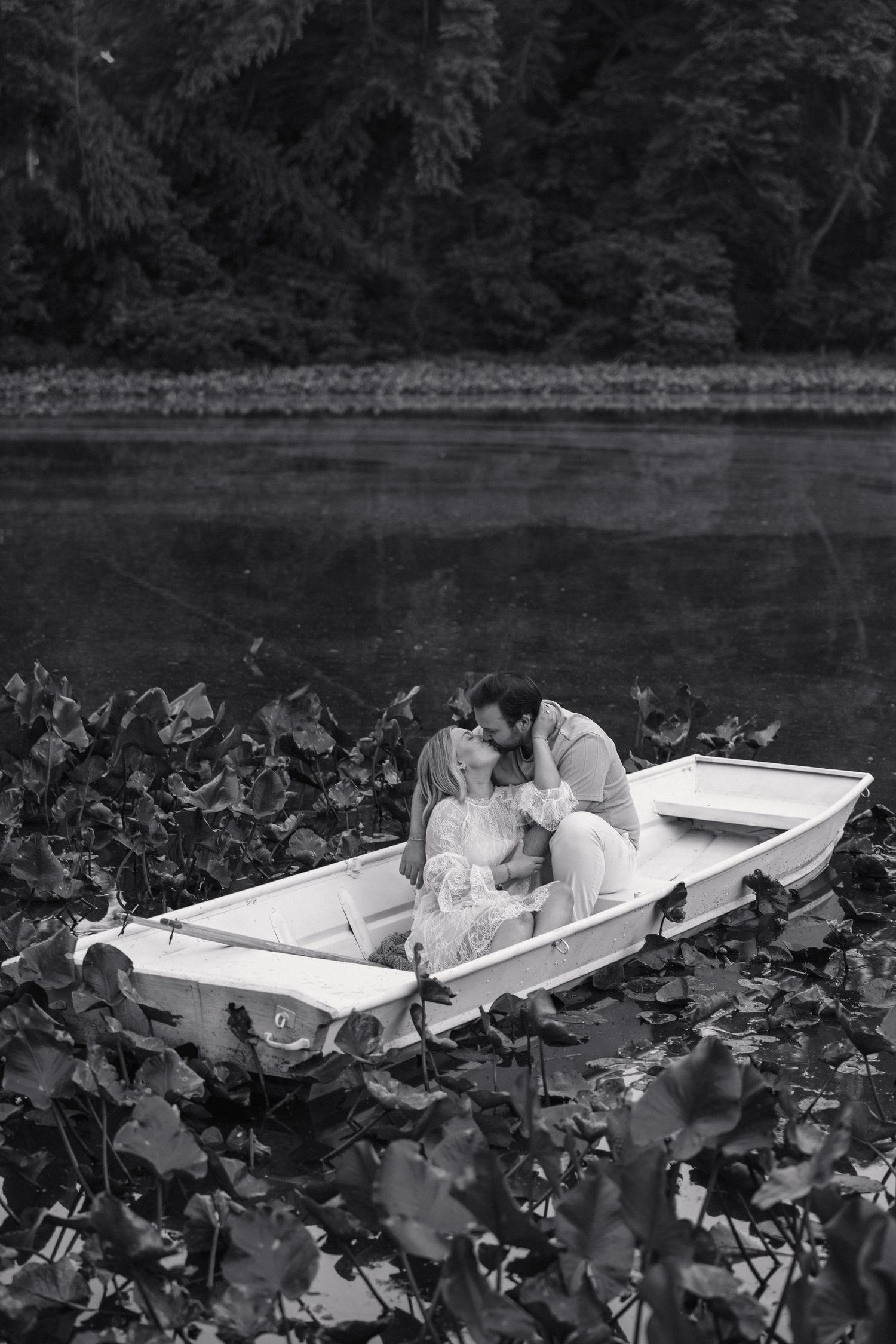 Black and white image of a couple sharing a tender kiss in a white rowboat surrounded by lily pads.