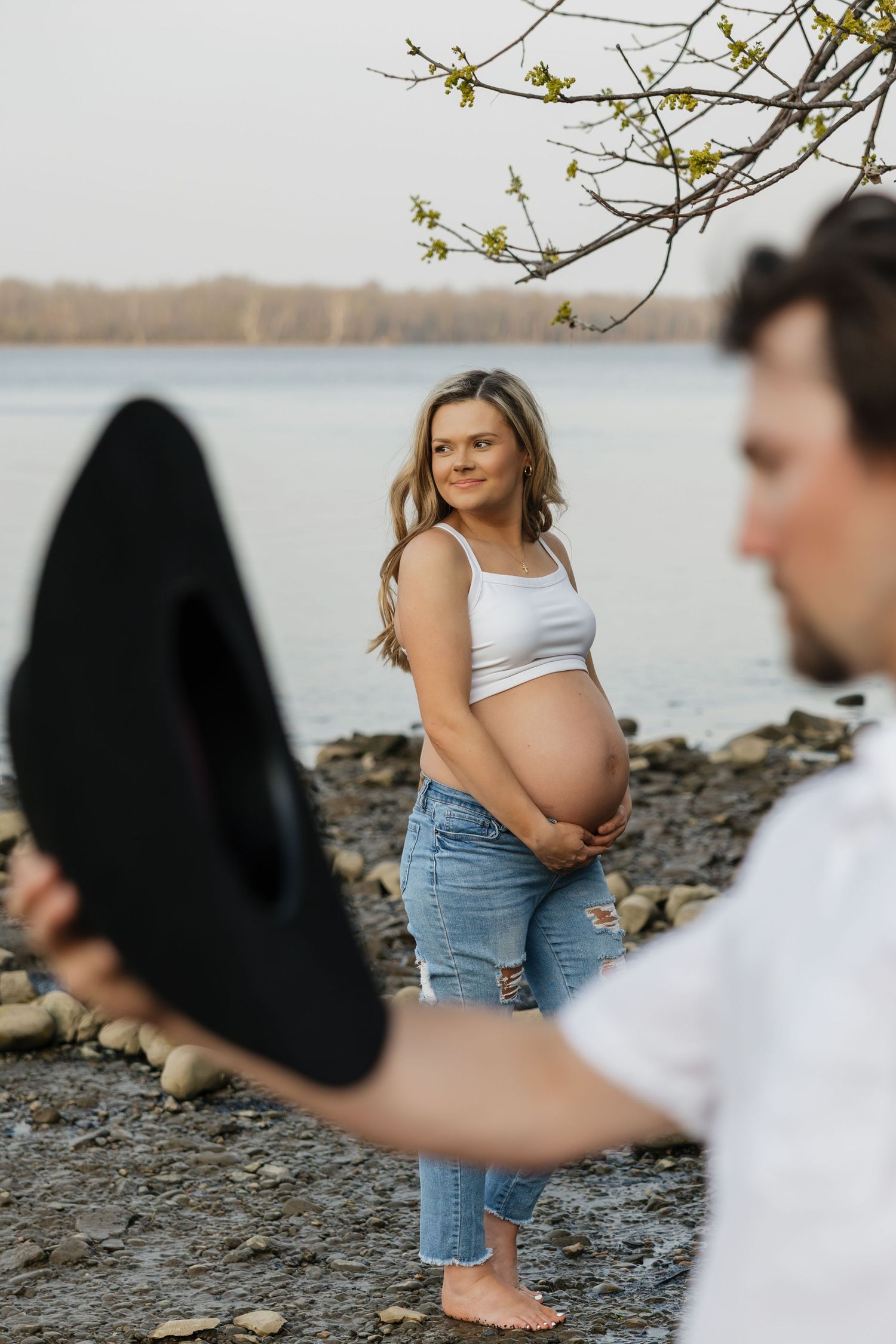 Smiling pregnant blonde woman cradles baby bump while man in white shirt holds black hat at rocky shore.