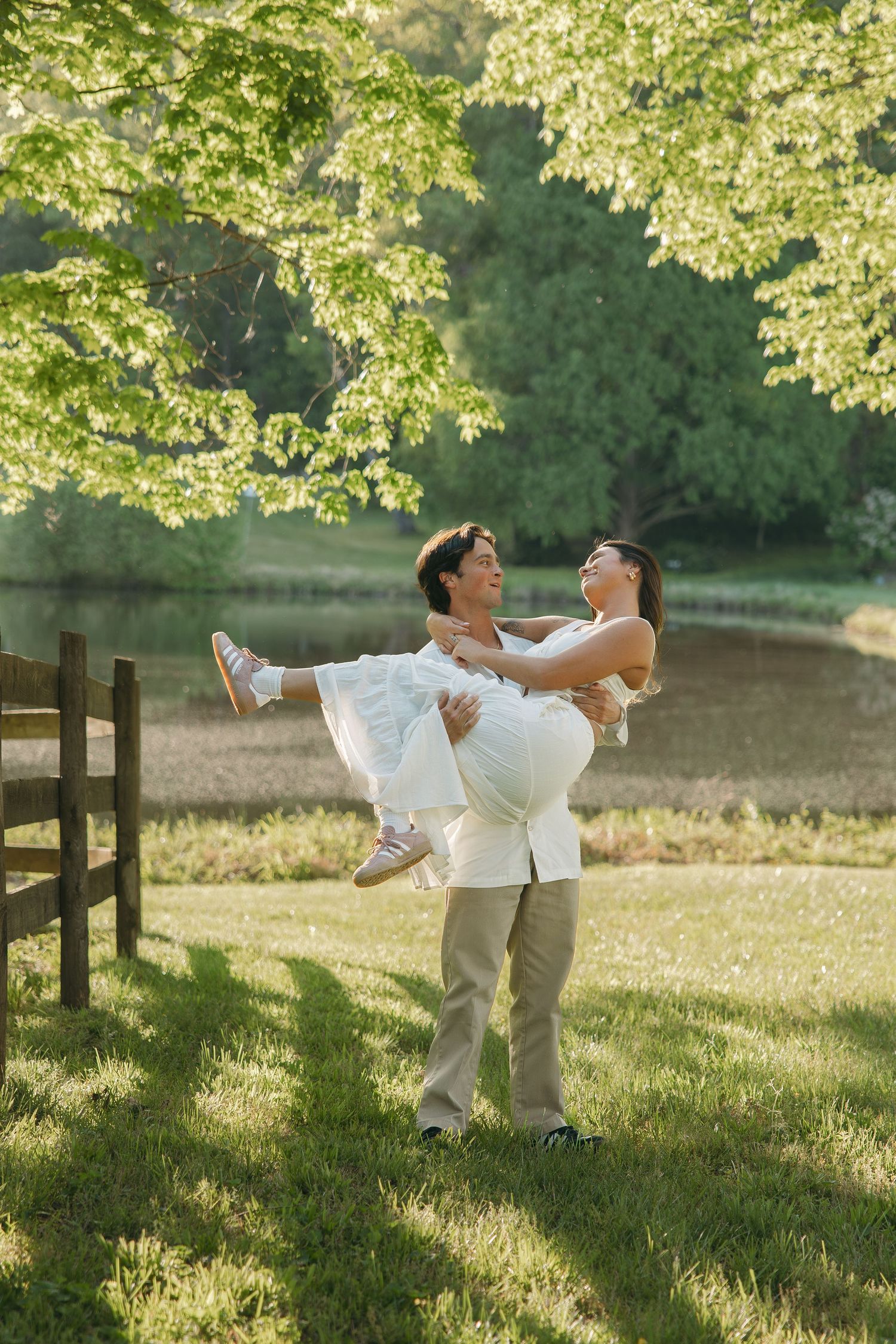 Man joyfully carrying woman in white dress in a sunny green park near a peaceful pond.