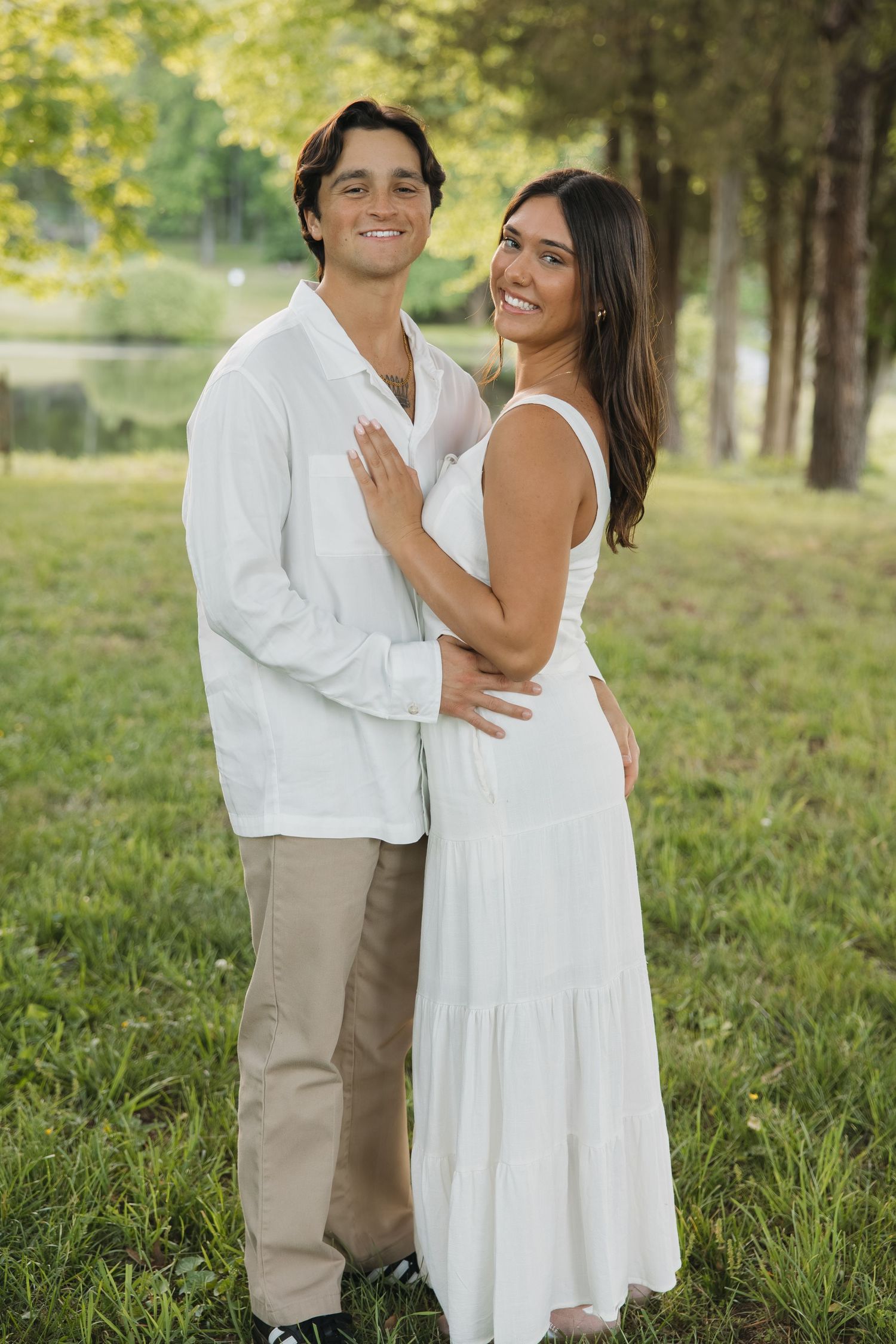 A smiling couple poses together on a green lawn surrounded by trees in warm sunlight.