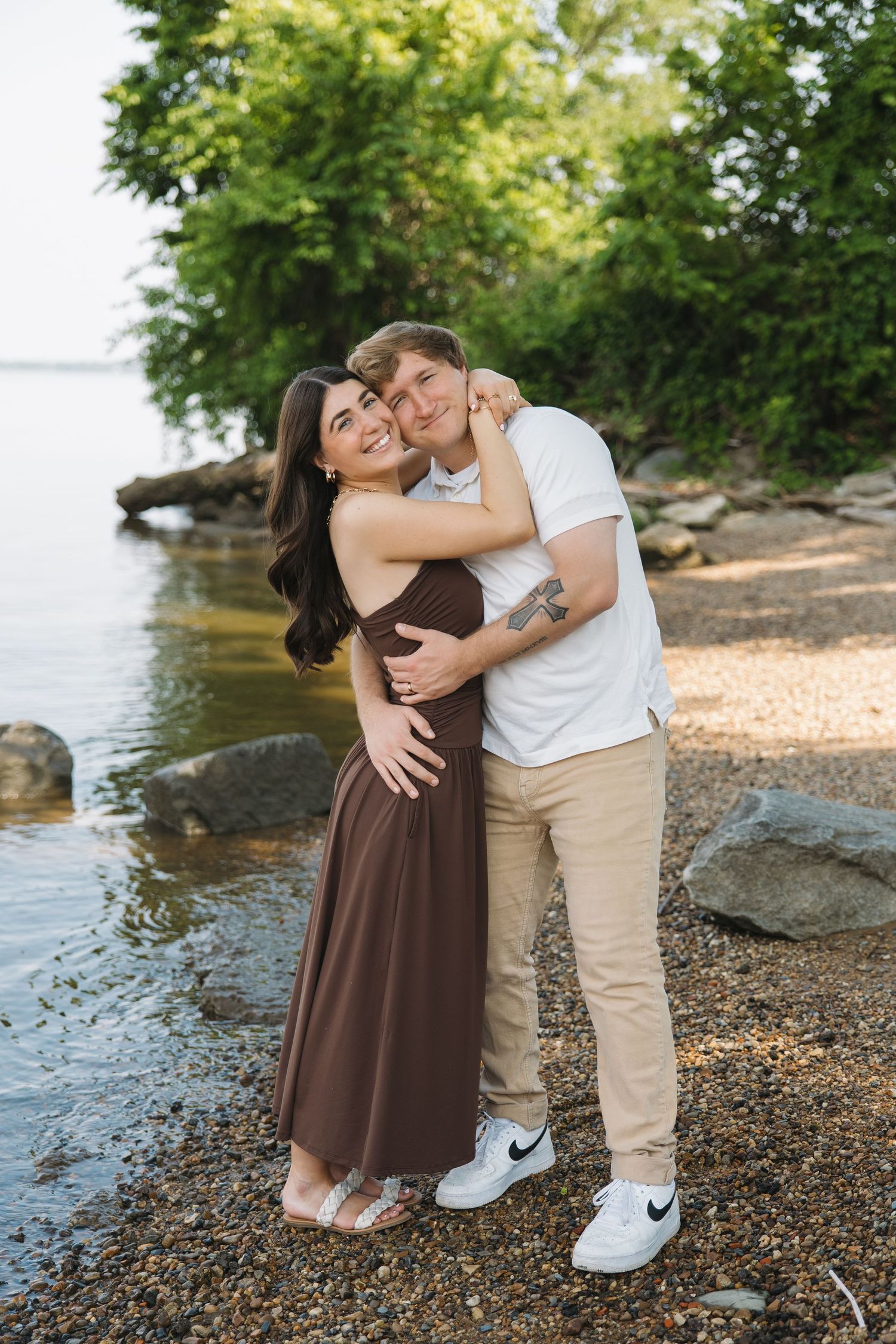 A couple shares a warm embrace while standing on a rocky shoreline surrounded by lush green trees.