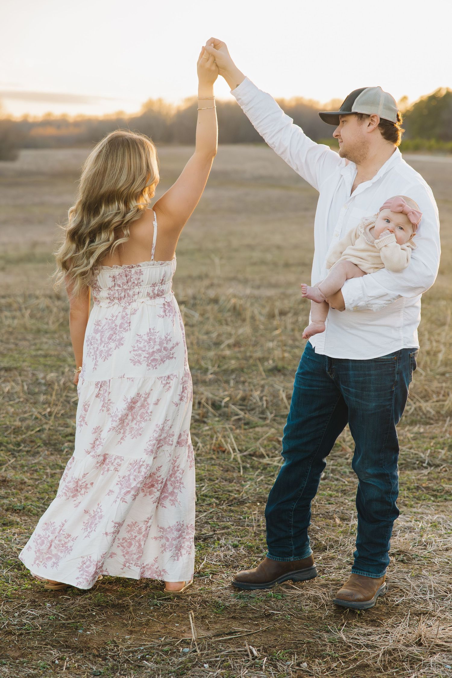 Father holding baby while dancing with mother in floral dress during golden hour in open field.