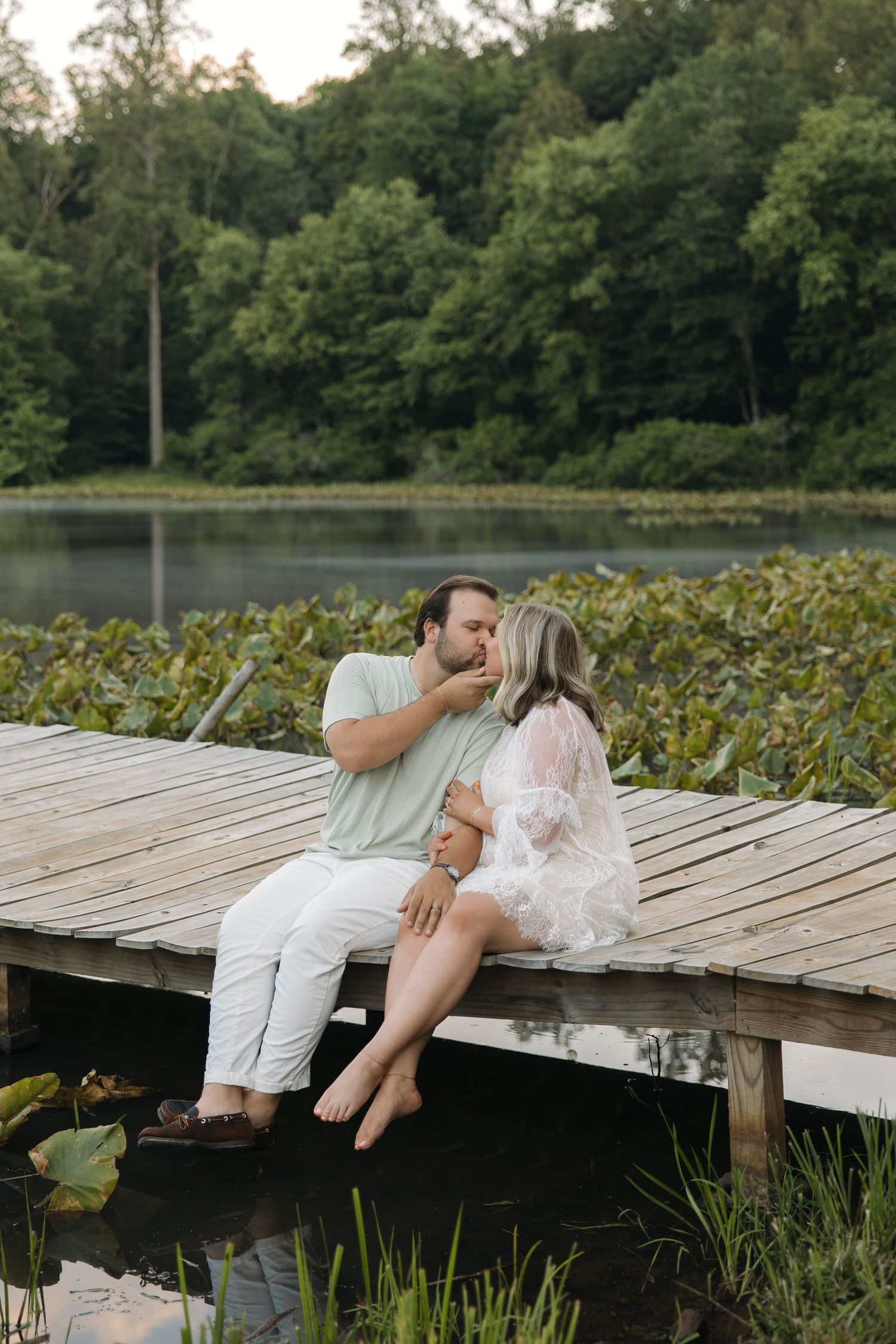 Couple sharing a tender kiss while sitting on a wooden dock by a lily pad lake with green trees behind.