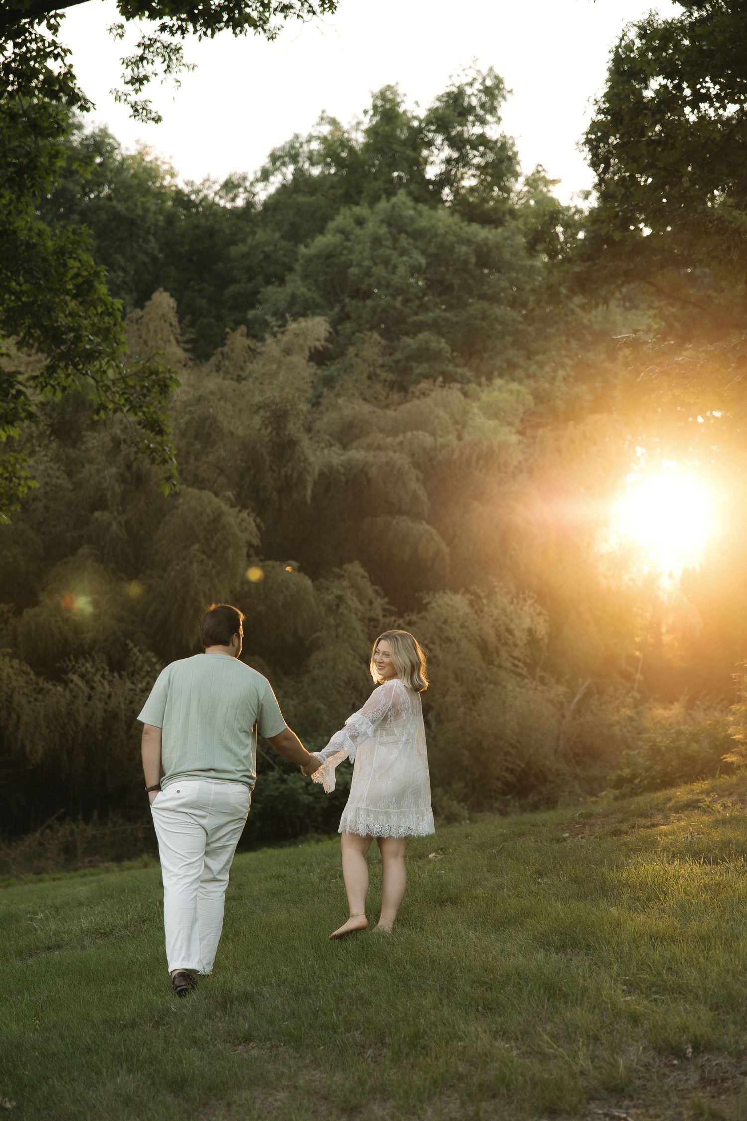Couple holding hands walking barefoot through green grass toward glowing golden sunset through lush trees.