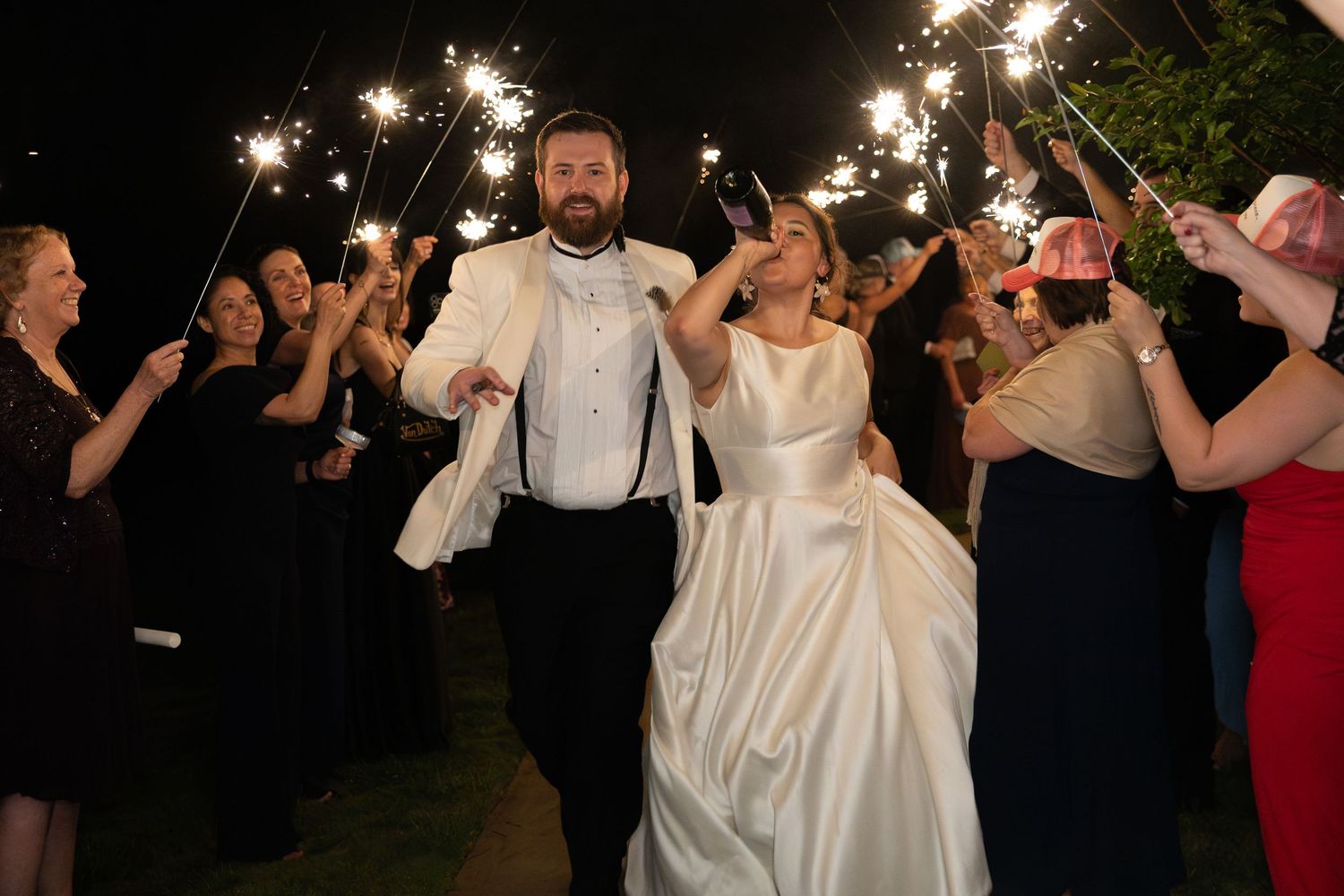 Smiling bride and groom walk through sparkler exit tunnel with wedding guests celebrating their reception send-off at night.