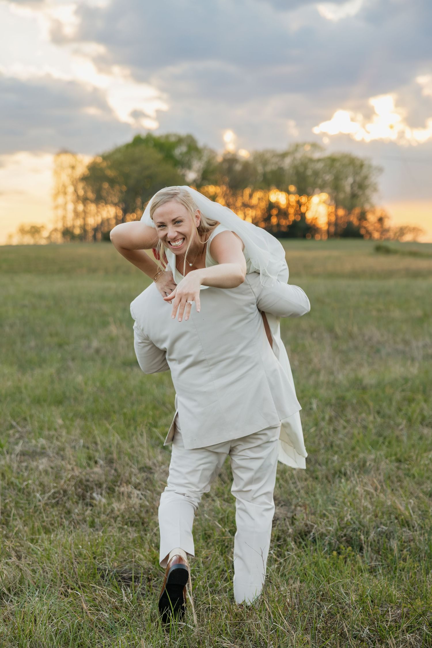 Laughing bride carried on grooms back in a sunlit field at sunset.