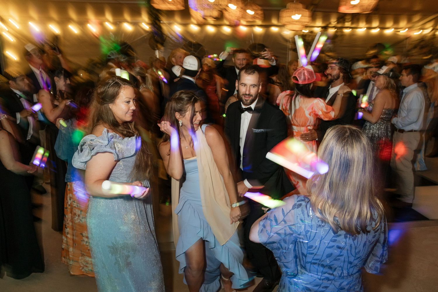 Wedding reception dance floor with guests in formal attire dancing under warm string lights with LED glow sticks.