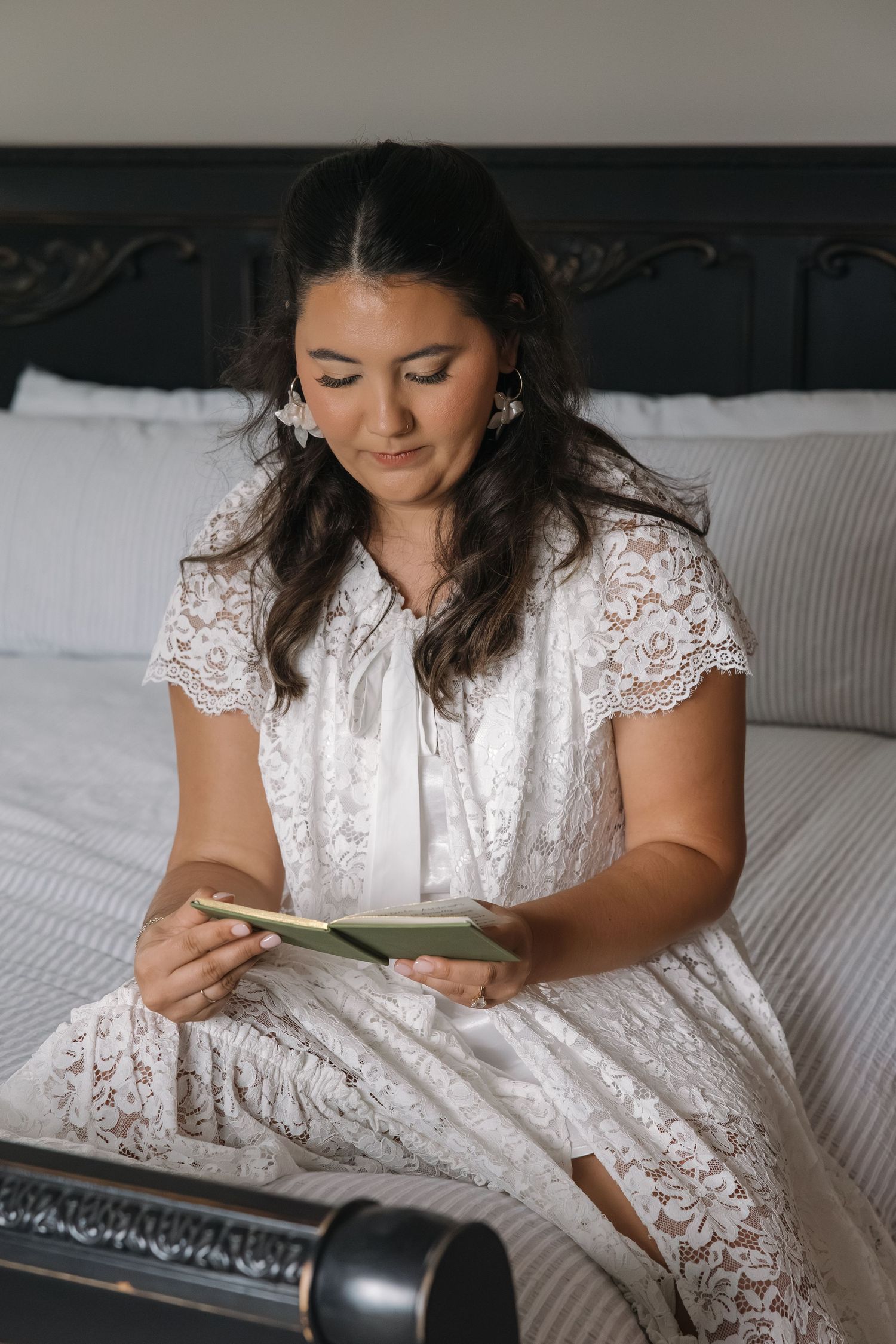 Bride in white lace dress reading a card while sitting on a bed before her wedding.