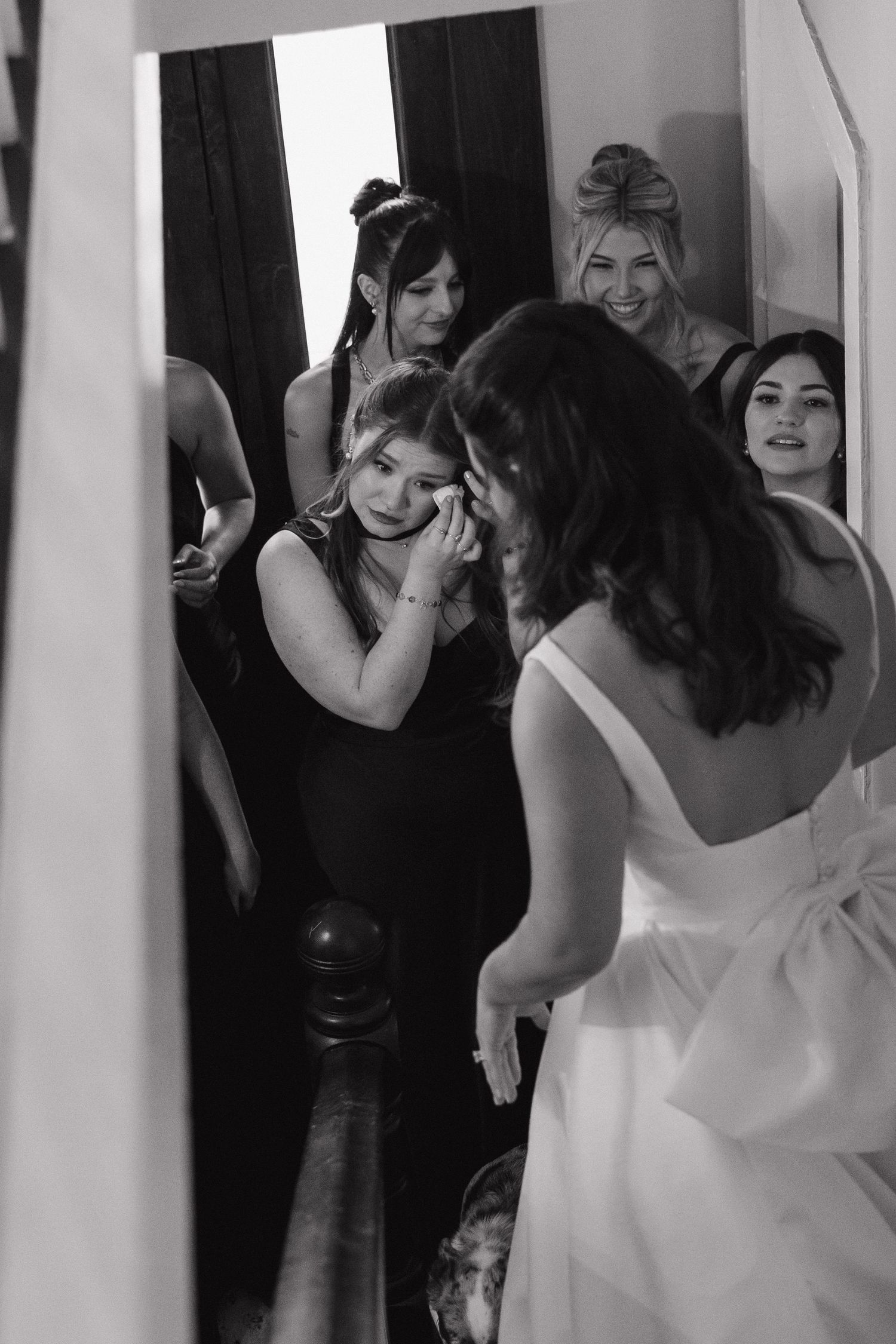 Emotional bridesmaid wipes tears seeing bride in wedding gown surrounded by smiling bridesmaids.