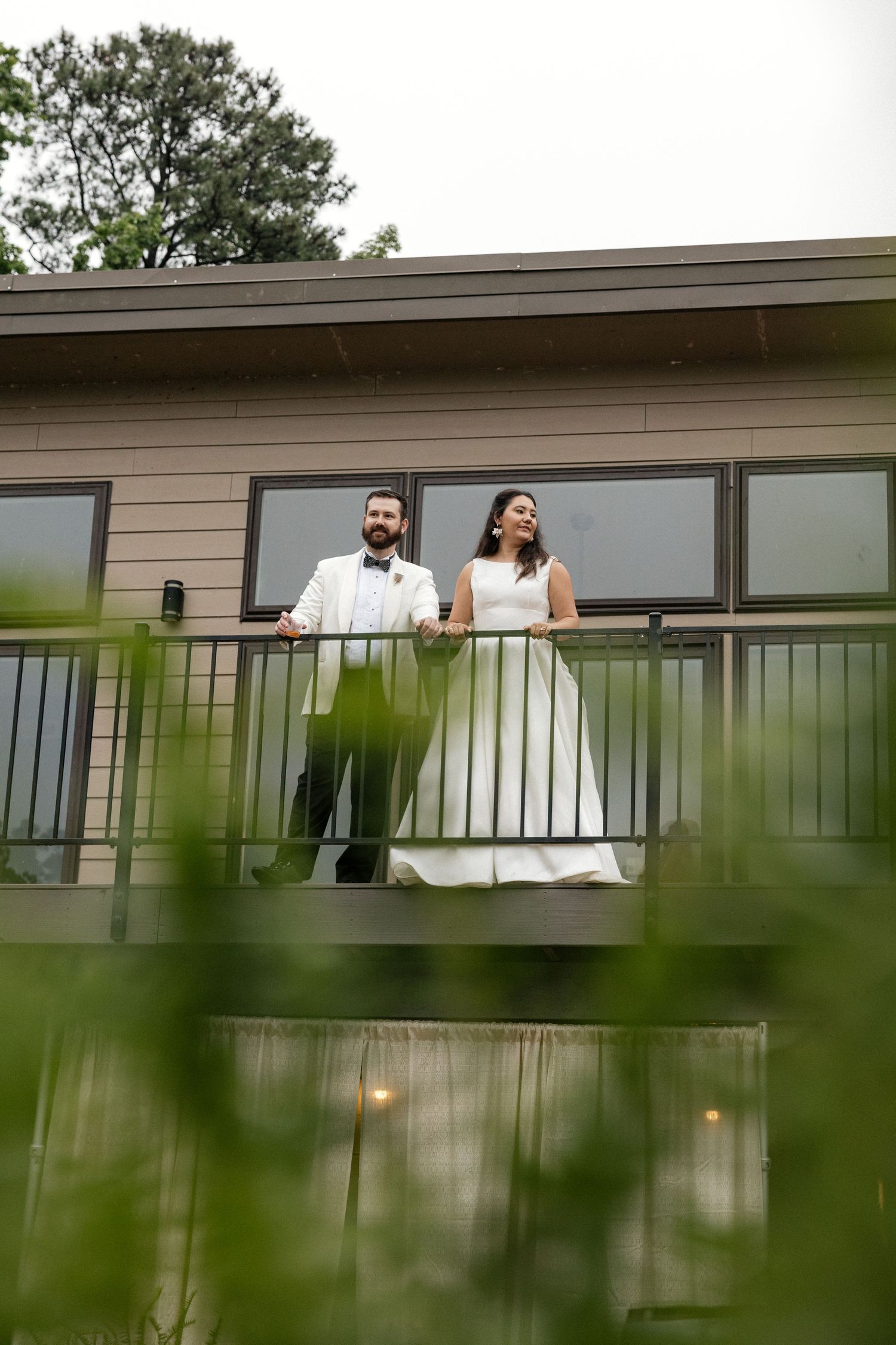 Bride and groom posing on a balcony overlooking lush greenery through tall grass foreground.