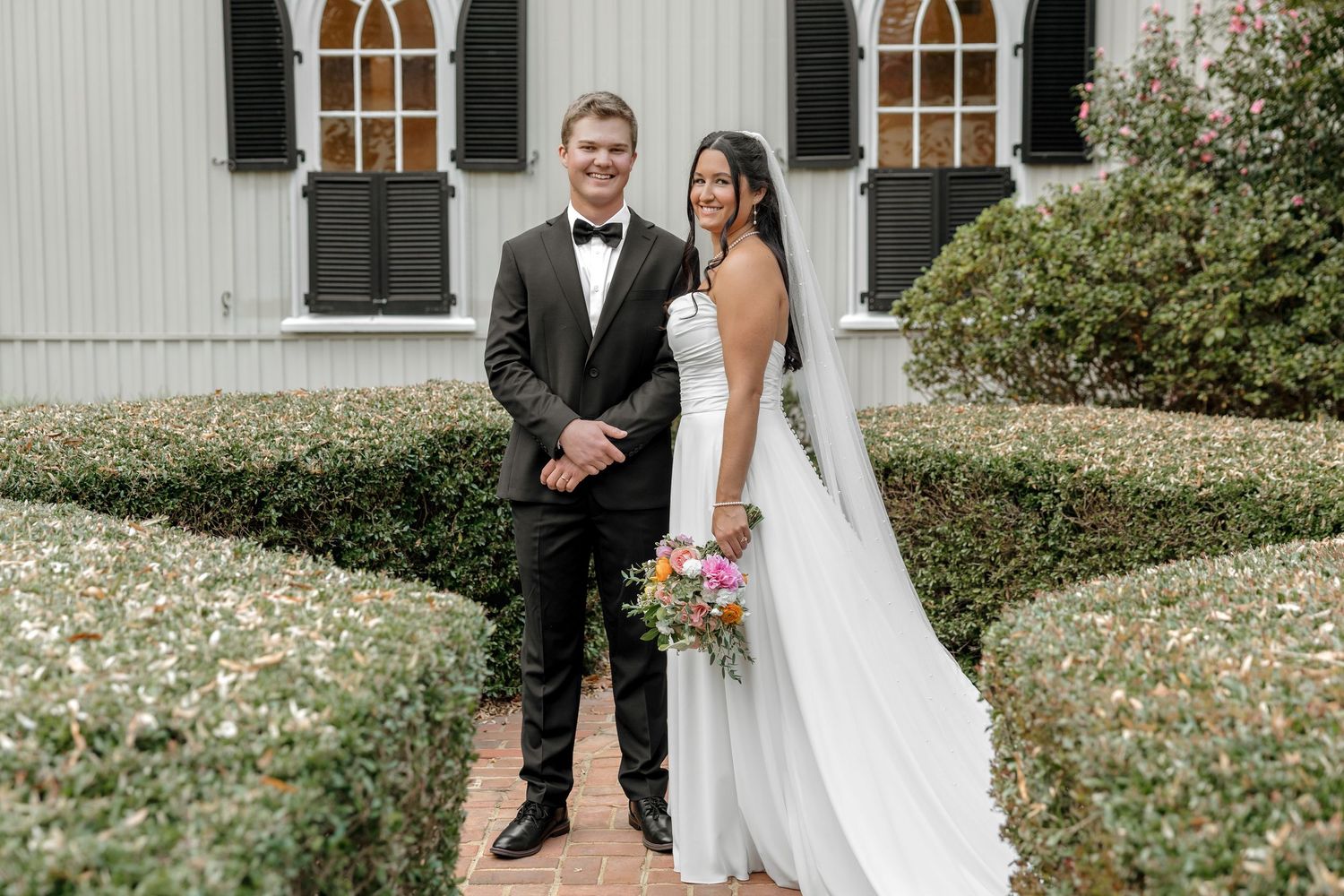 Bride and groom posing together outside a white church surrounded by manicured hedges on their wedding day.