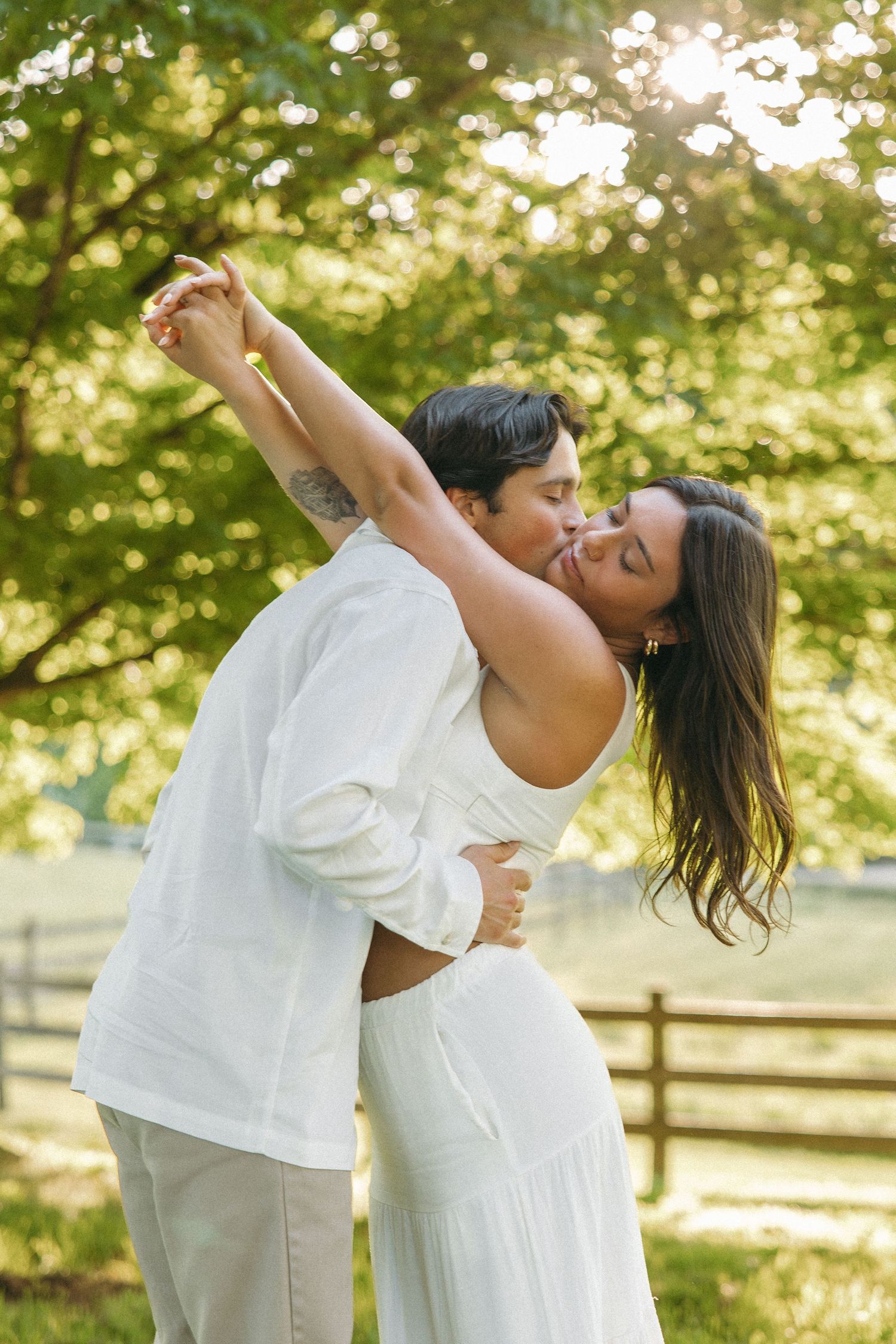 Couple sharing a romantic dance outdoors, kissing under golden sunlight with lush green trees and wooden fence behind.