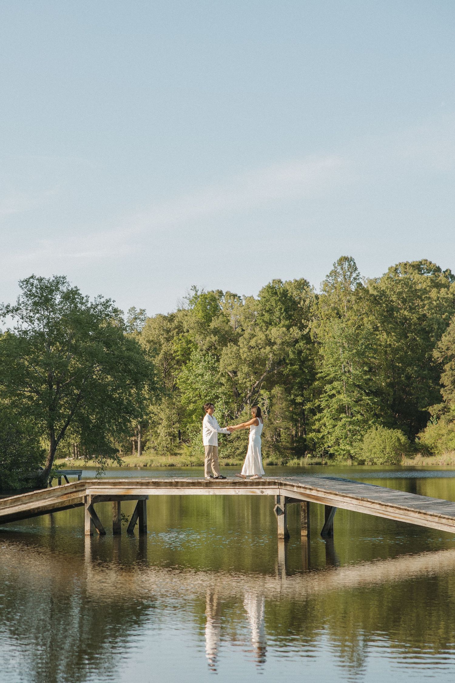 Couple holding hands on a wooden dock over a calm lake surrounded by lush green trees on a sunny day.