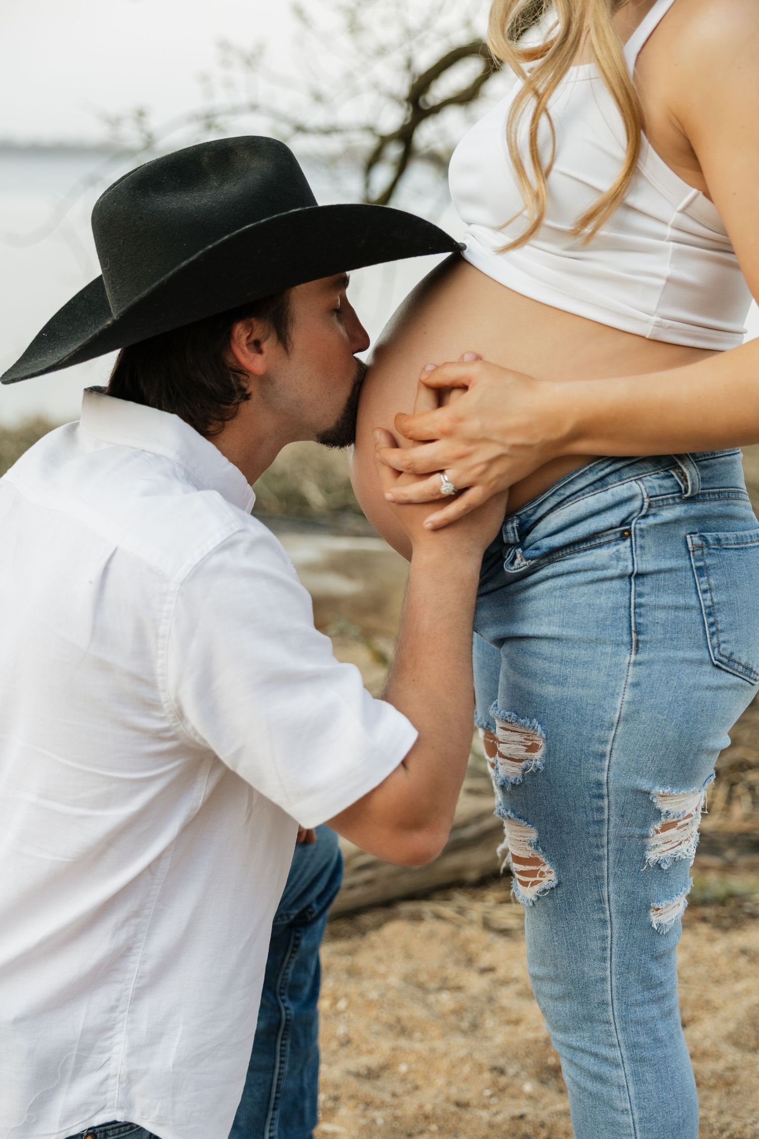 Man in black cowboy hat kneeling to kiss pregnant woman's bare baby bump near riverside.