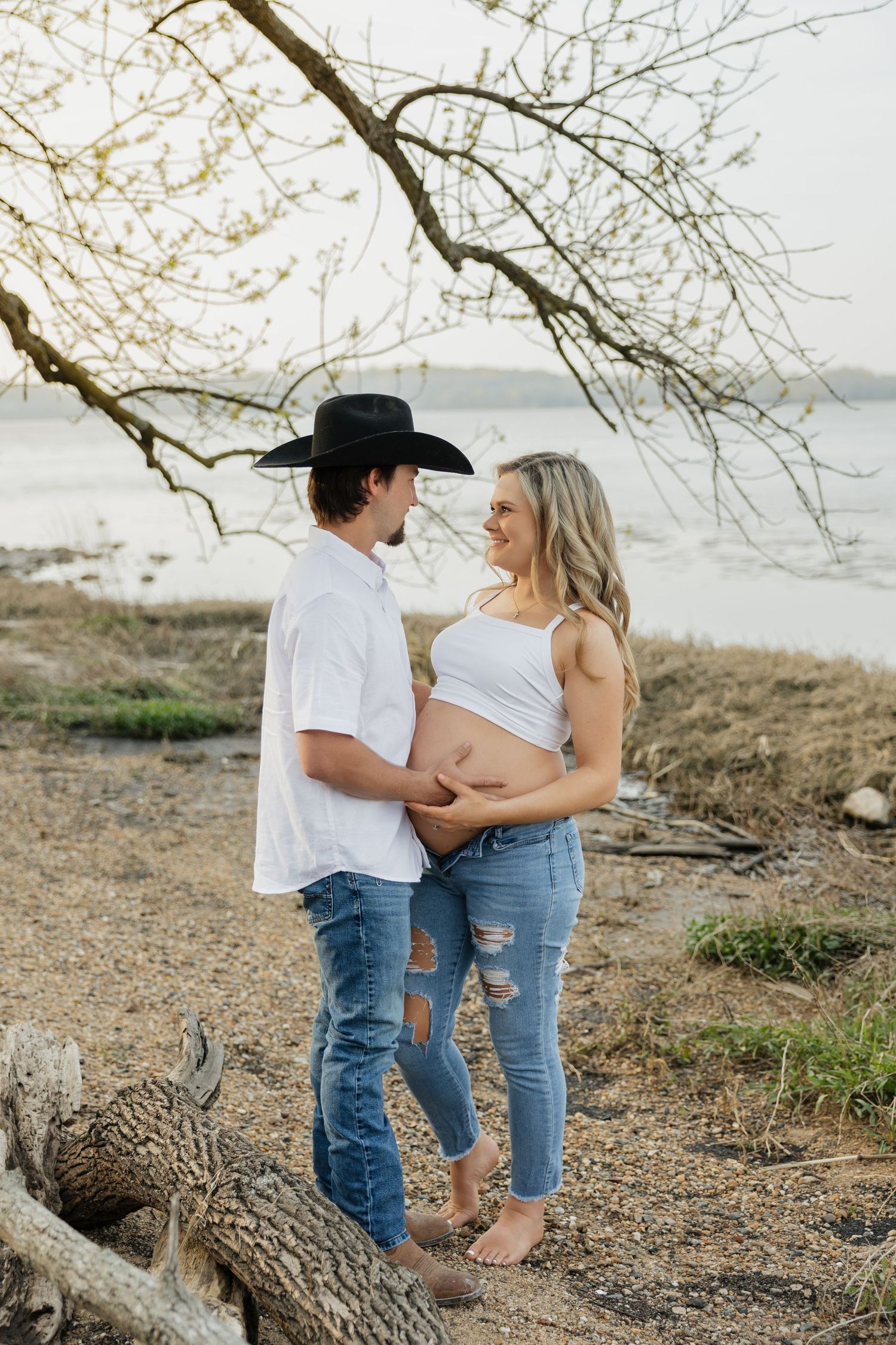 Expecting couple holding baby bump and smiling at each other under a tree near a lake shoreline.