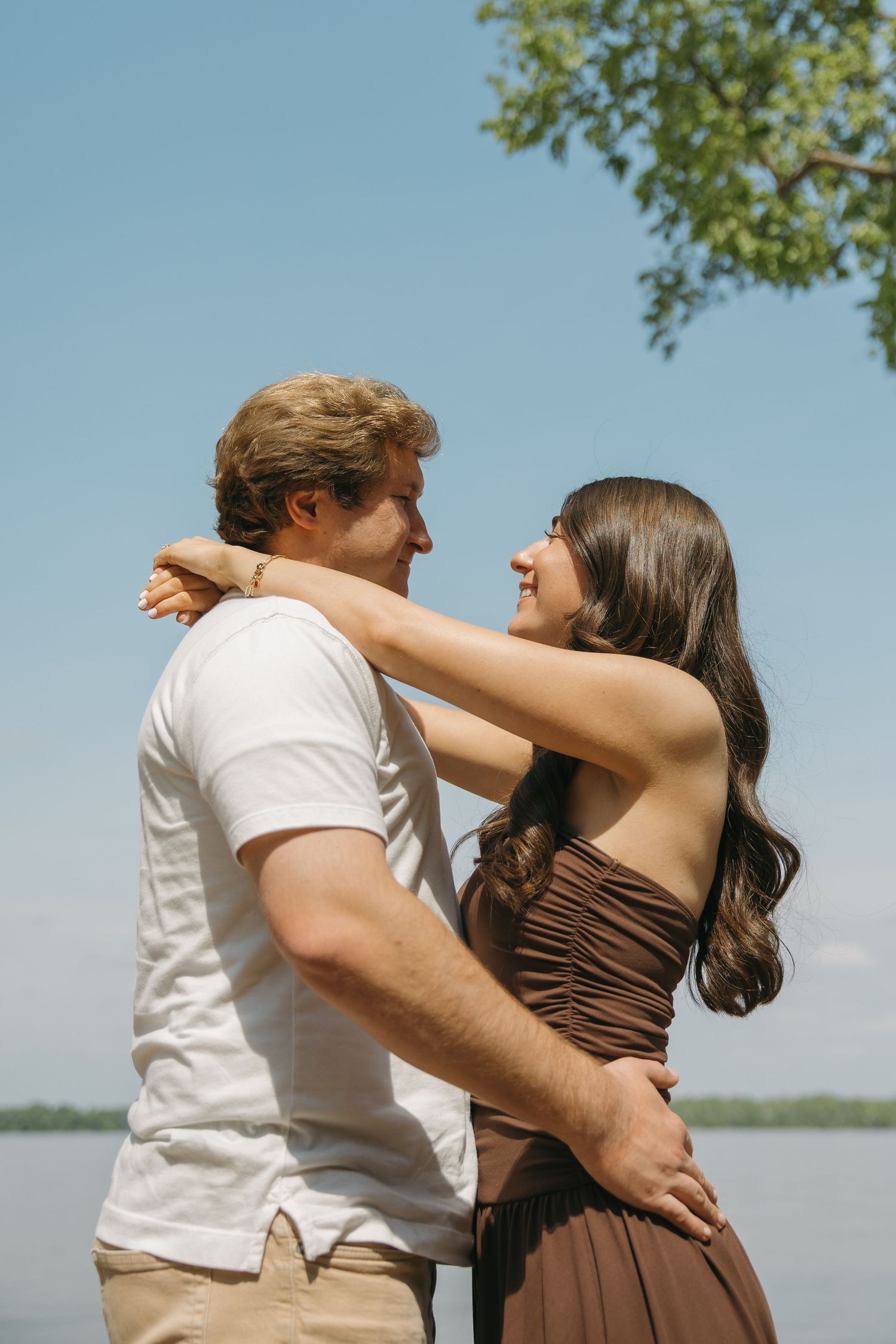 Couple embracing and smiling at each other outdoors by a lake under a clear blue sky.