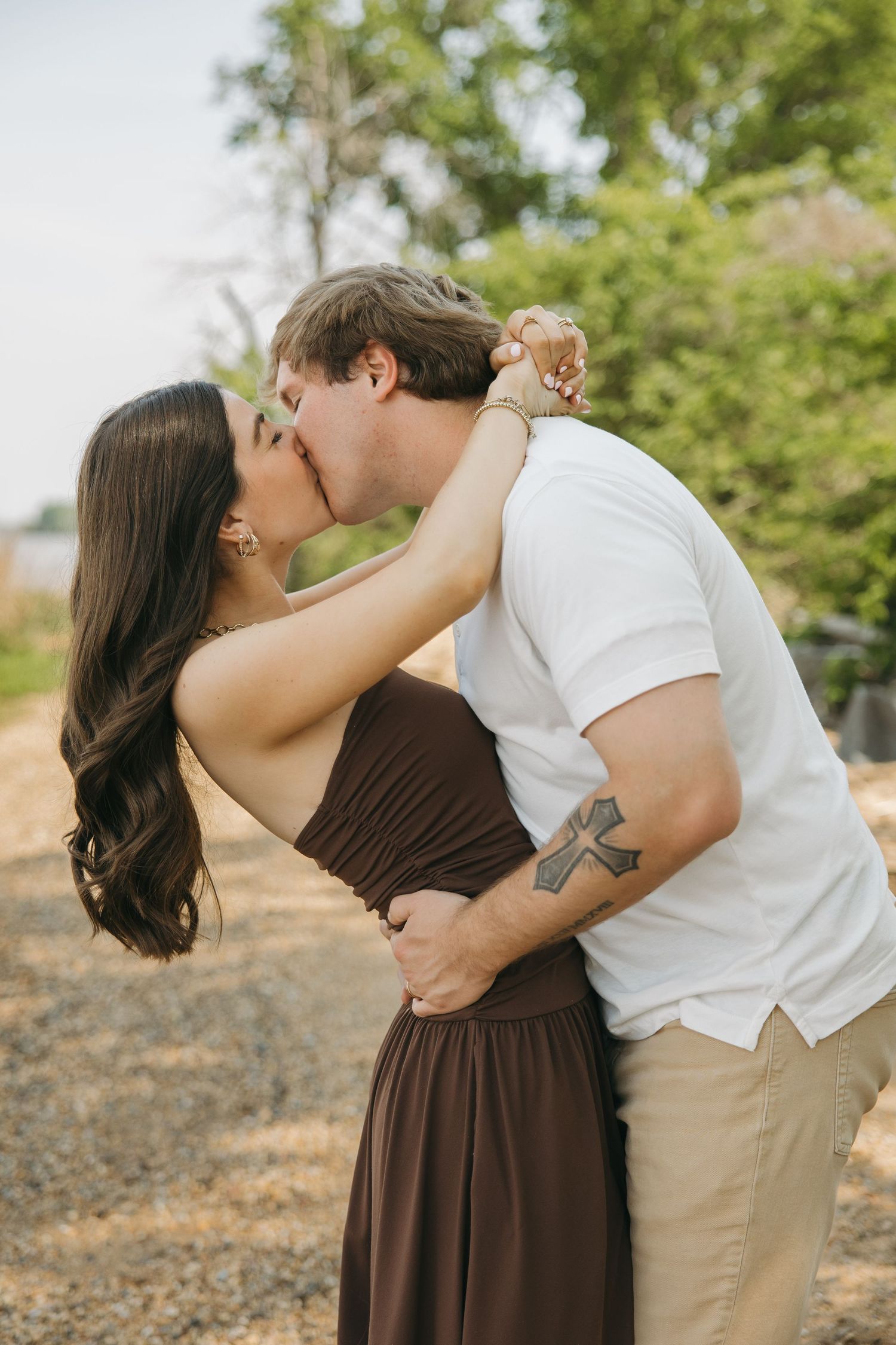 A couple shares a passionate kiss outdoors, woman in brown strapless dress pulling man close by the neck.