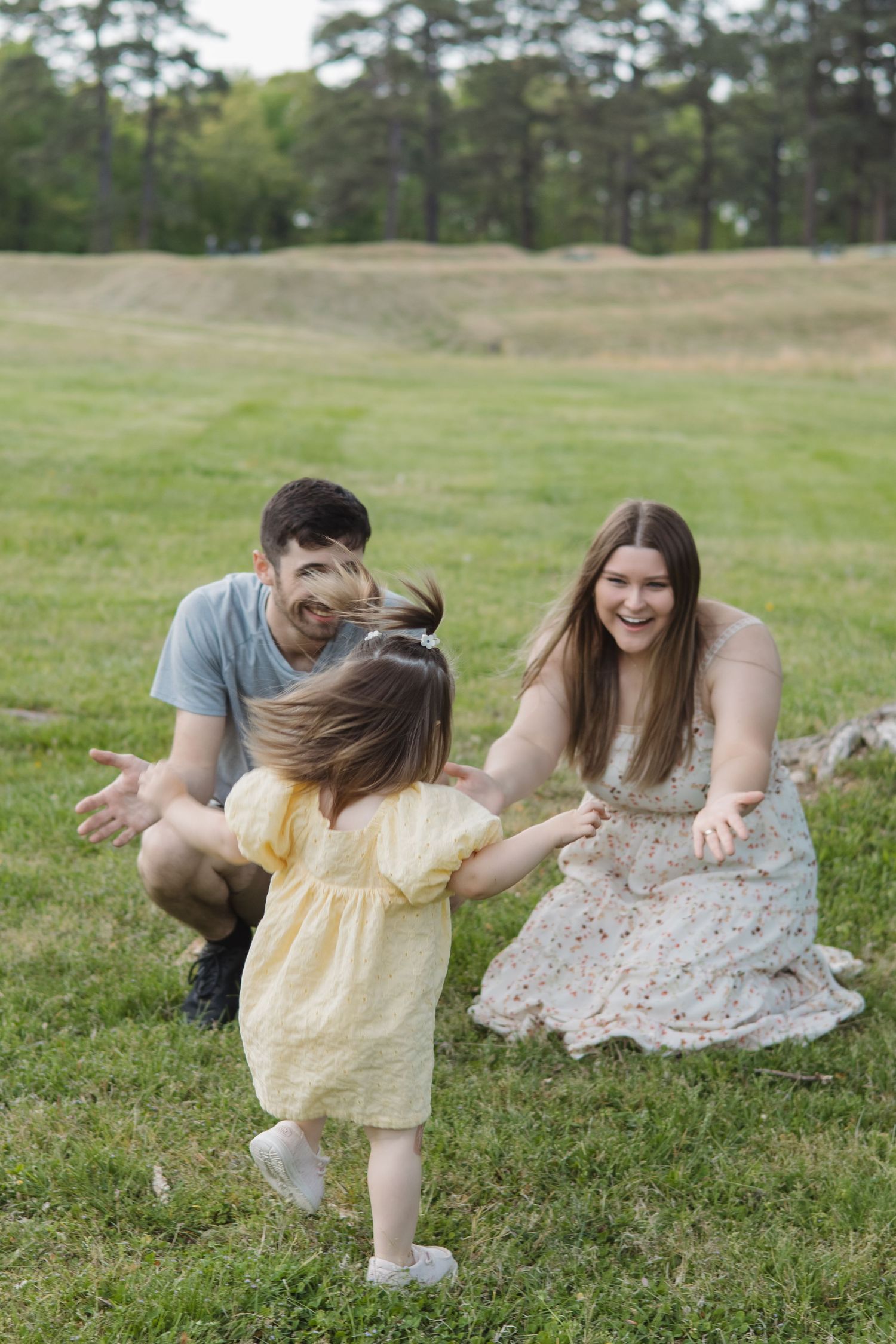 Joyful family moment with parents crouching on grass as their toddler daughter runs toward them with arms open.