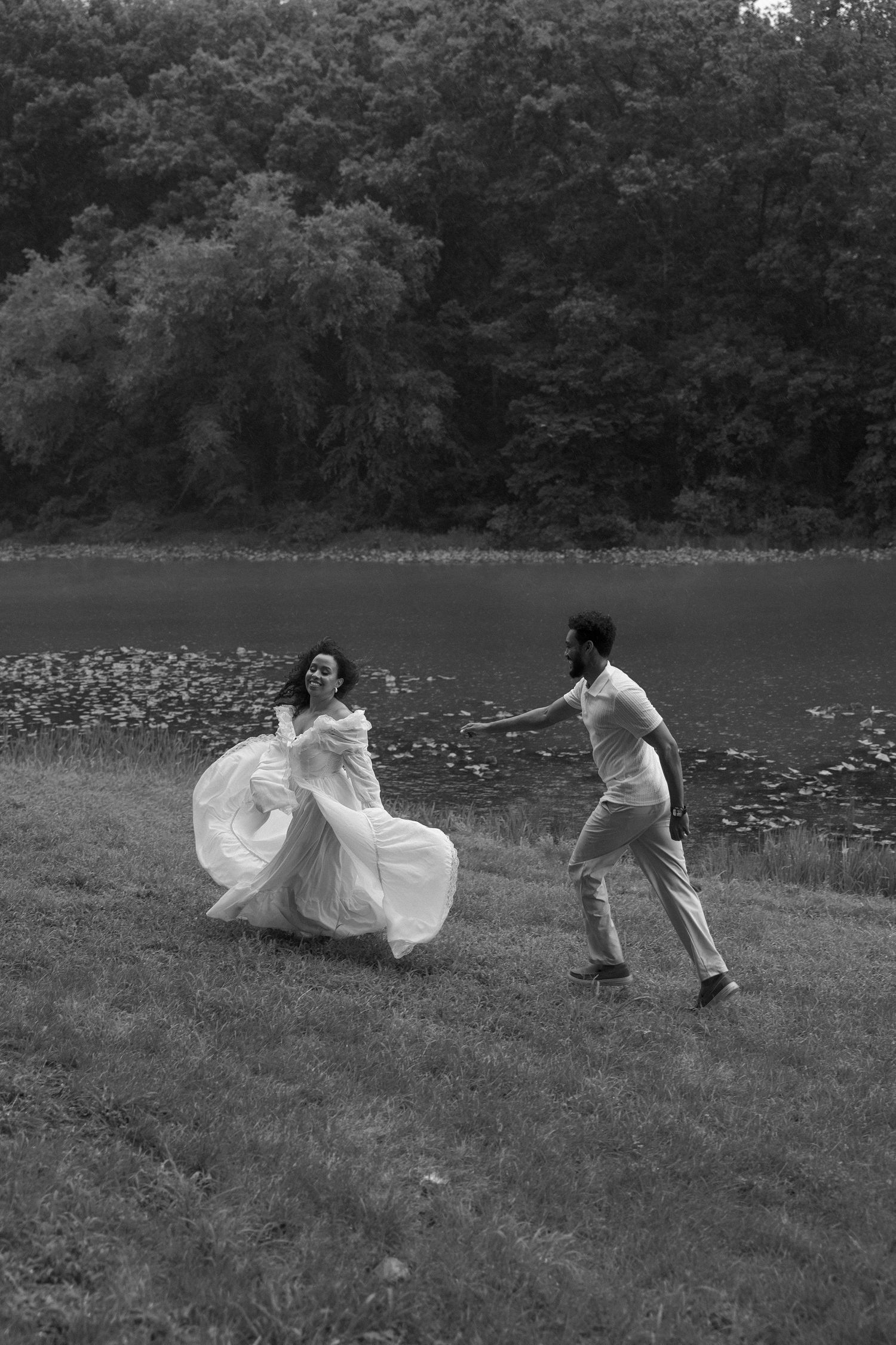 Black and white photo of a bride in flowing white gown running near a lake as a man reaches toward her.