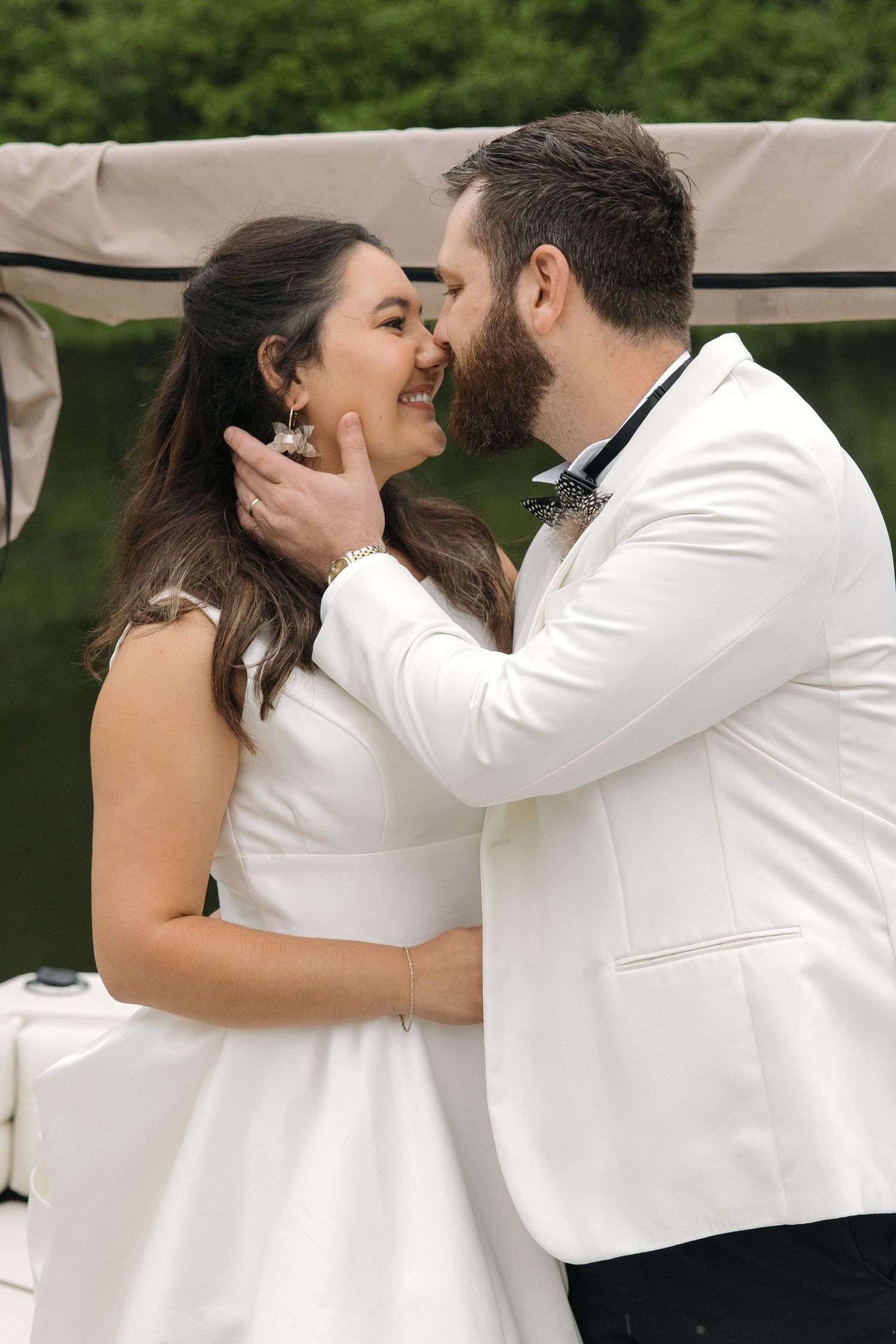 Groom in white tuxedo tenderly holding bride's face as they share a joyful moment on a boat.