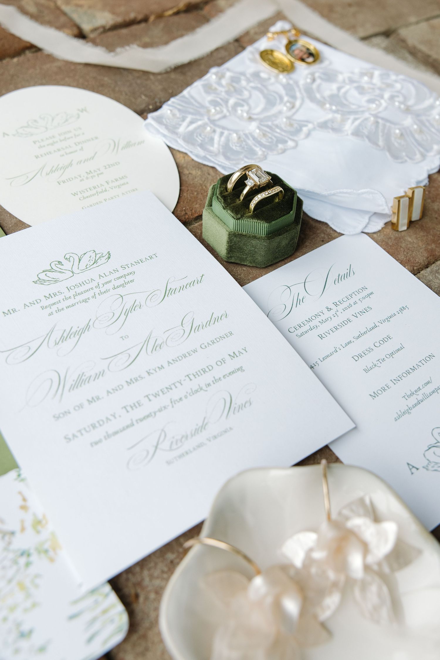 Elegant wedding invitation suite with green velvet ring box, gold rings, lace handkerchief and pearl earrings on brick.
