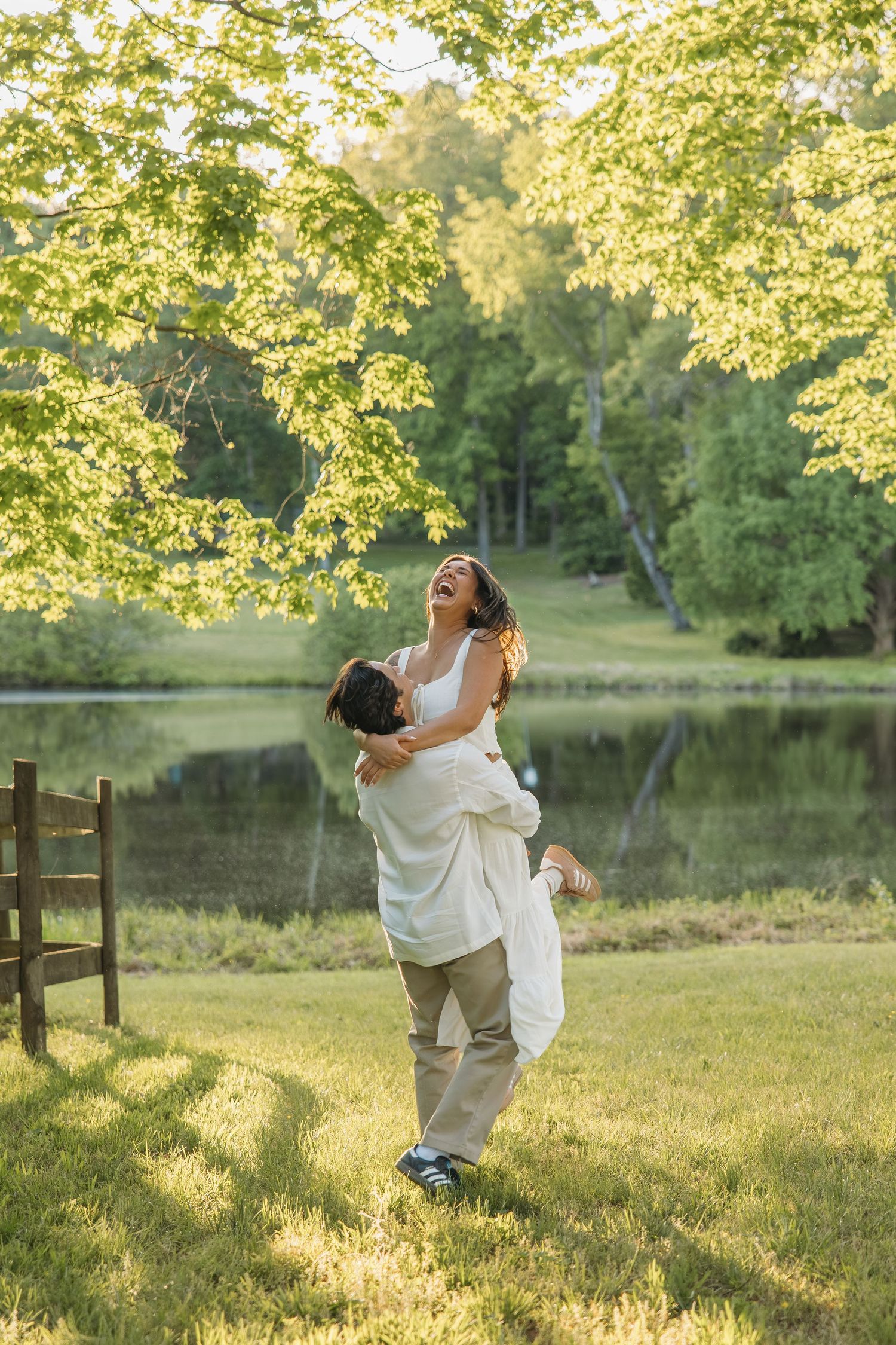 Groom lifting laughing bride in white dress outdoors near a pond with green trees in golden sunlight.