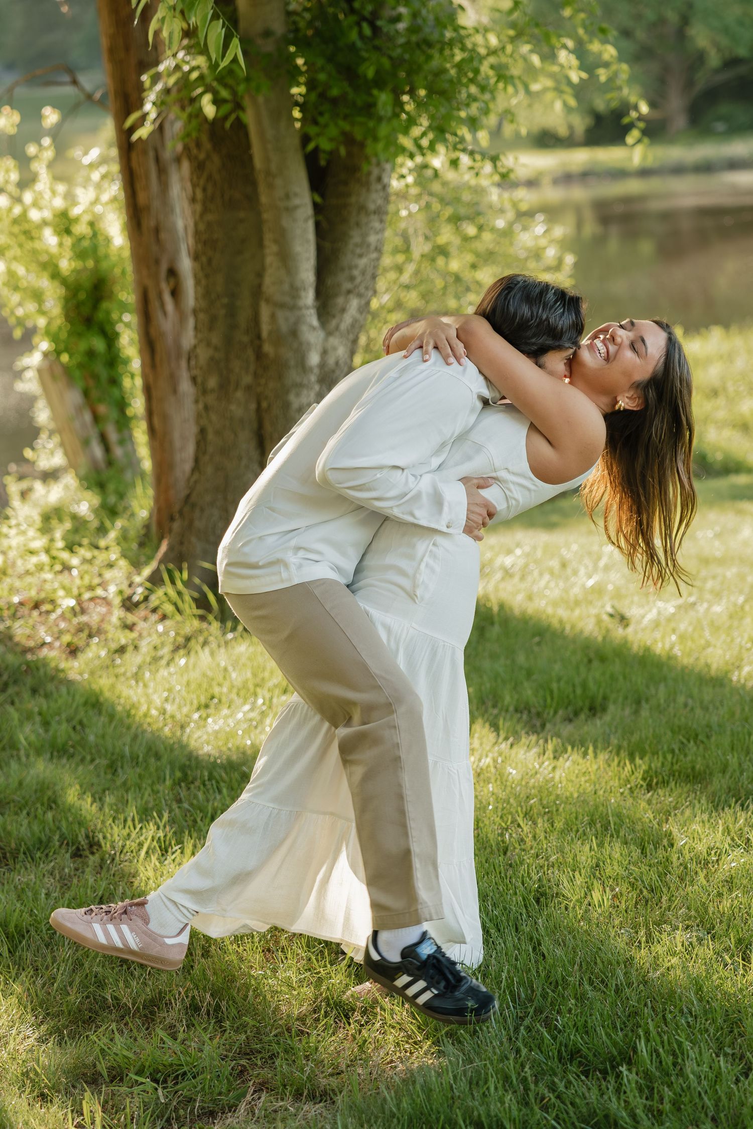 Couple laughing and embracing playfully near a tree in a sunlit green park by a tranquil lake.