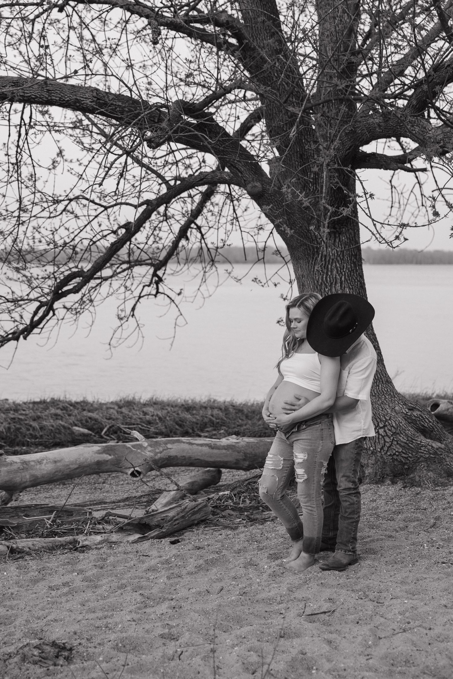 Black and white maternity photo of couple under bare tree by lake, man embracing pregnant woman tenderly.