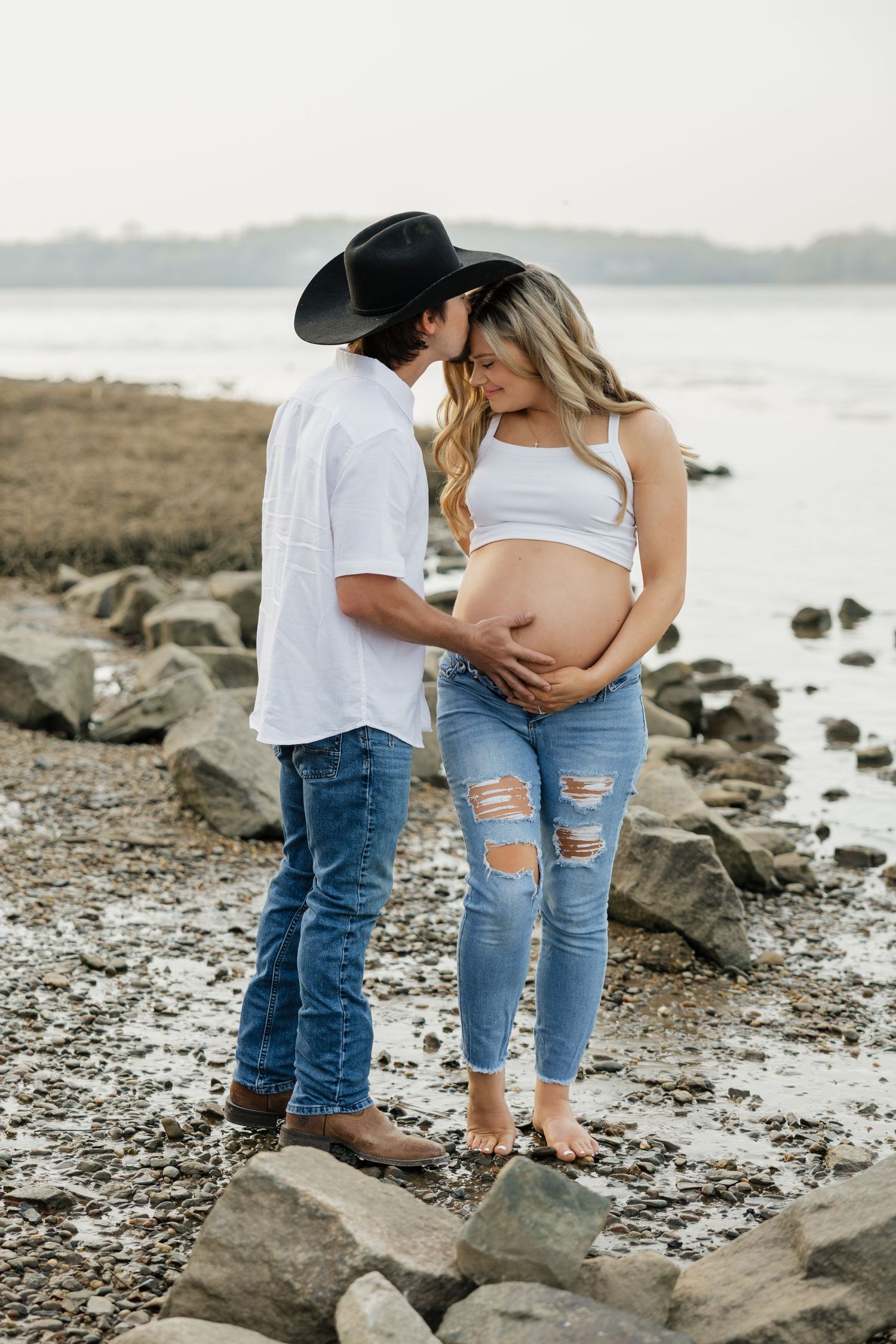 Couple sharing a kiss on rocky shoreline, pregnant woman in white crop top and ripped jeans near water.