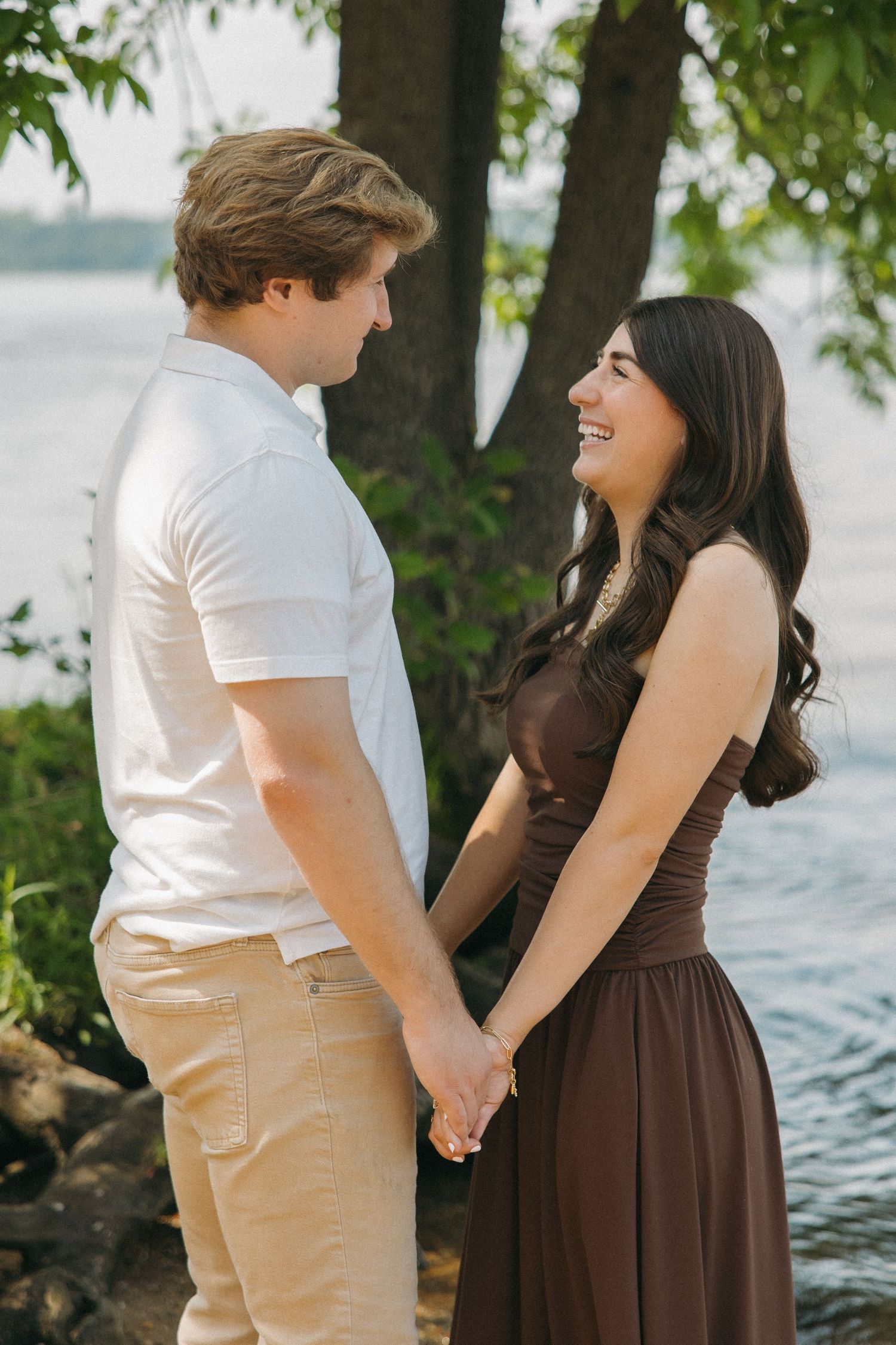A couple holds hands and smiles at each other near a tree by a peaceful lakeside.