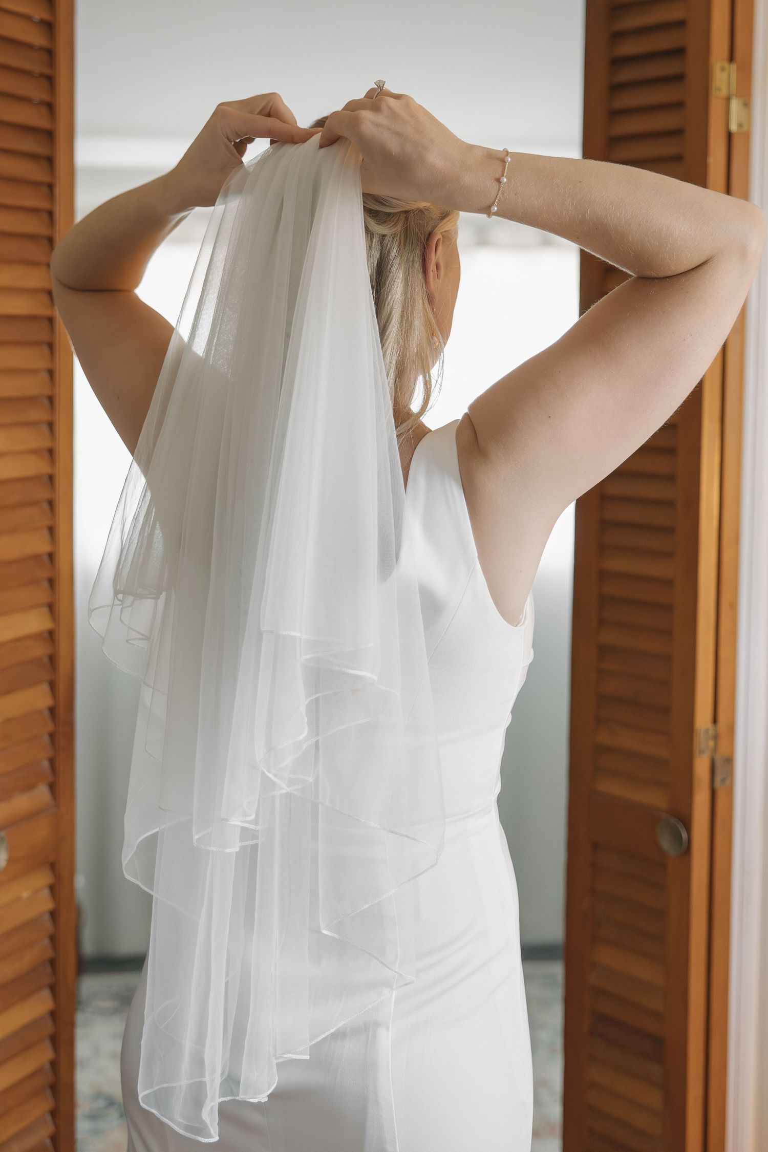 Bride in elegant white satin gown placing veil in hair, viewed from behind near wooden doors.