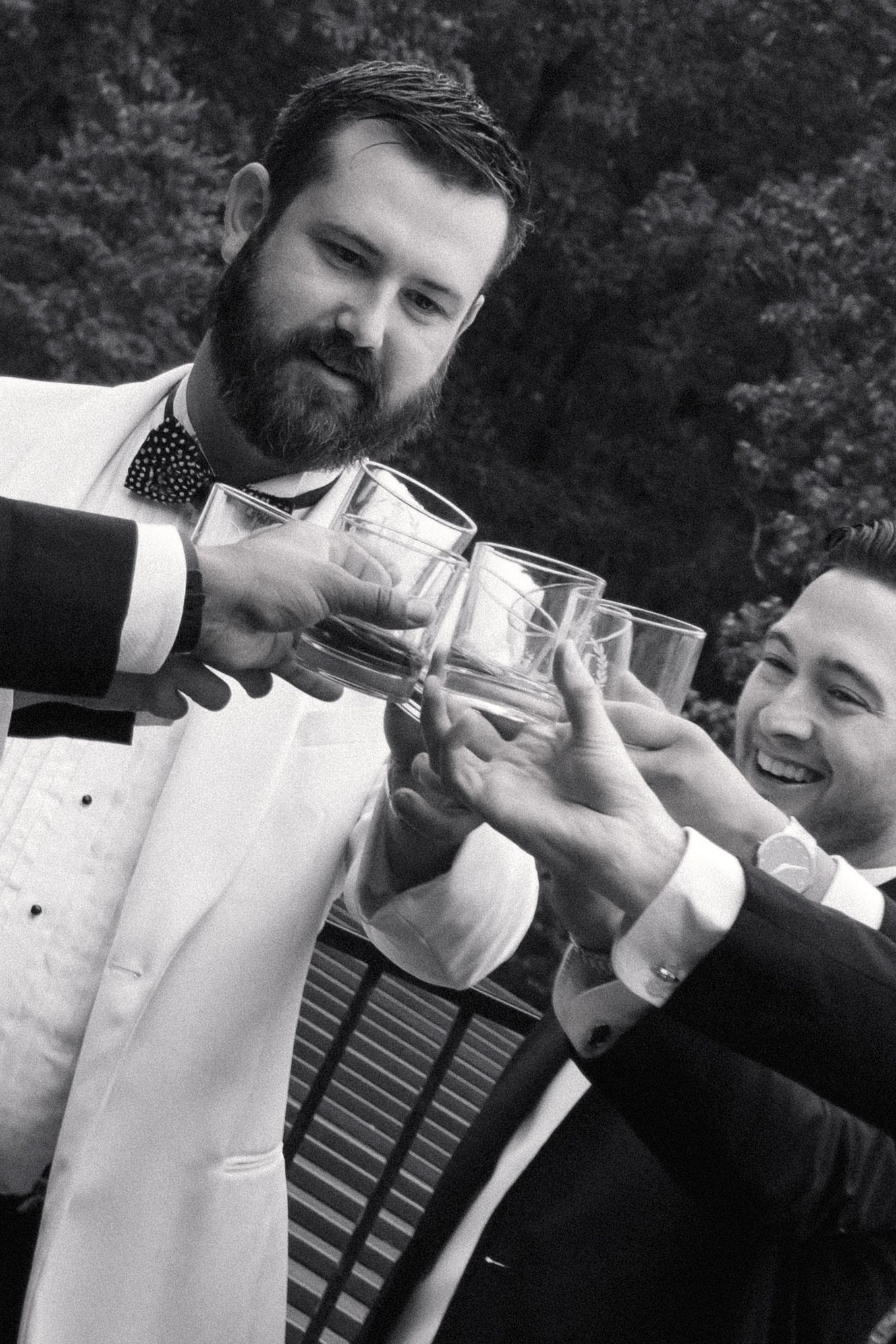 Black and white photo of bearded groom in white jacket toasting glasses with groomsmen at wedding celebration.