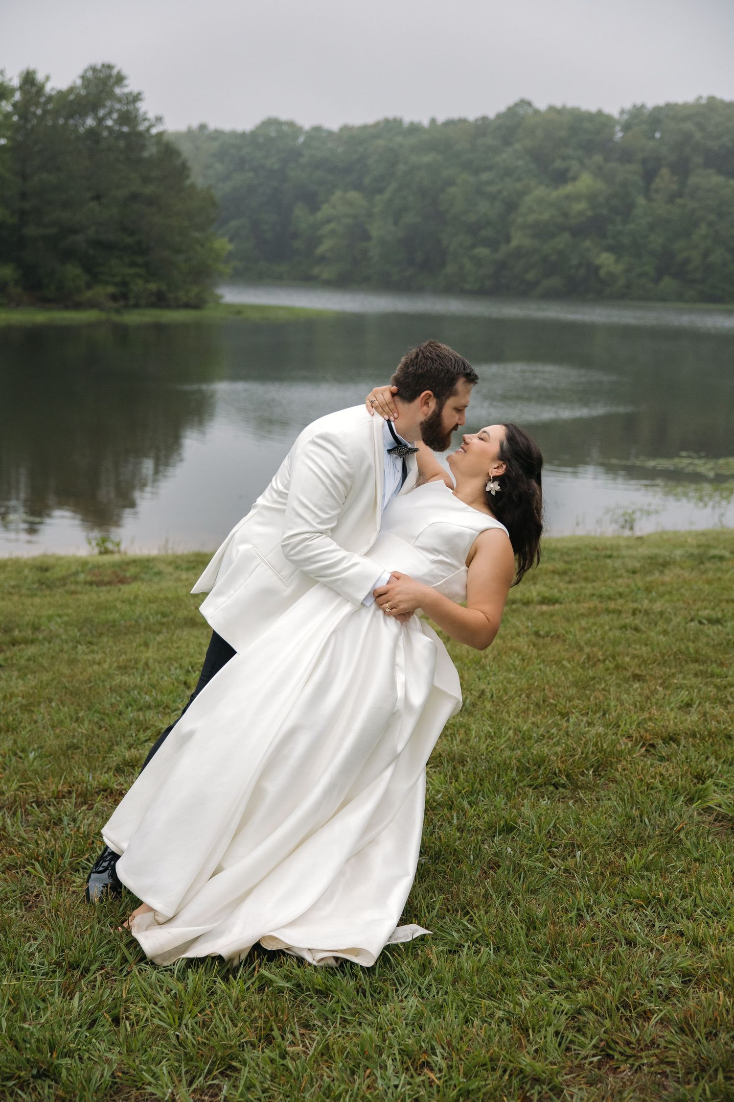 Groom dips bride in romantic pose near serene lake with lush green trees in background.