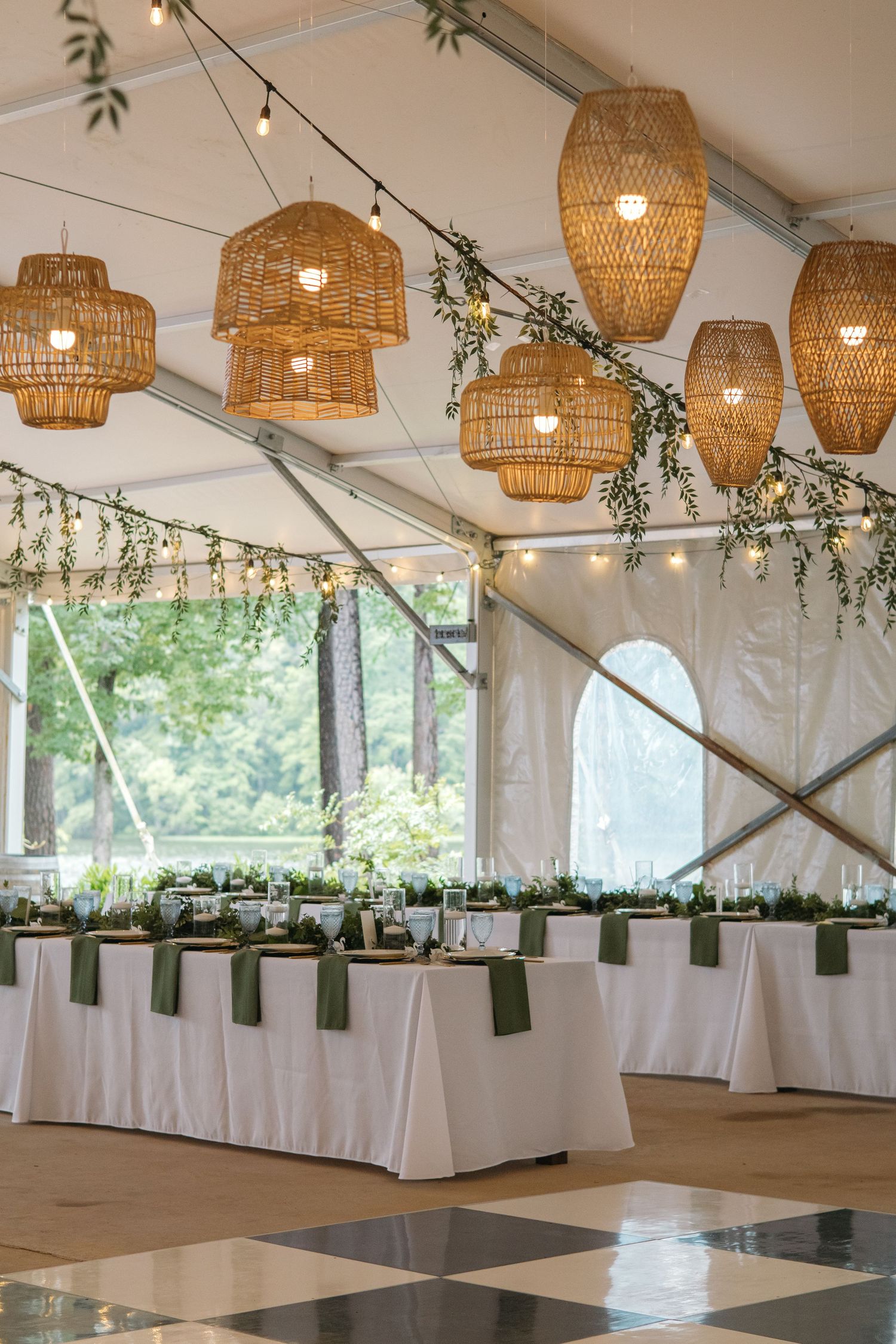 Wedding reception tent with wicker pendant lights, string lights, white tables, and green napkins.