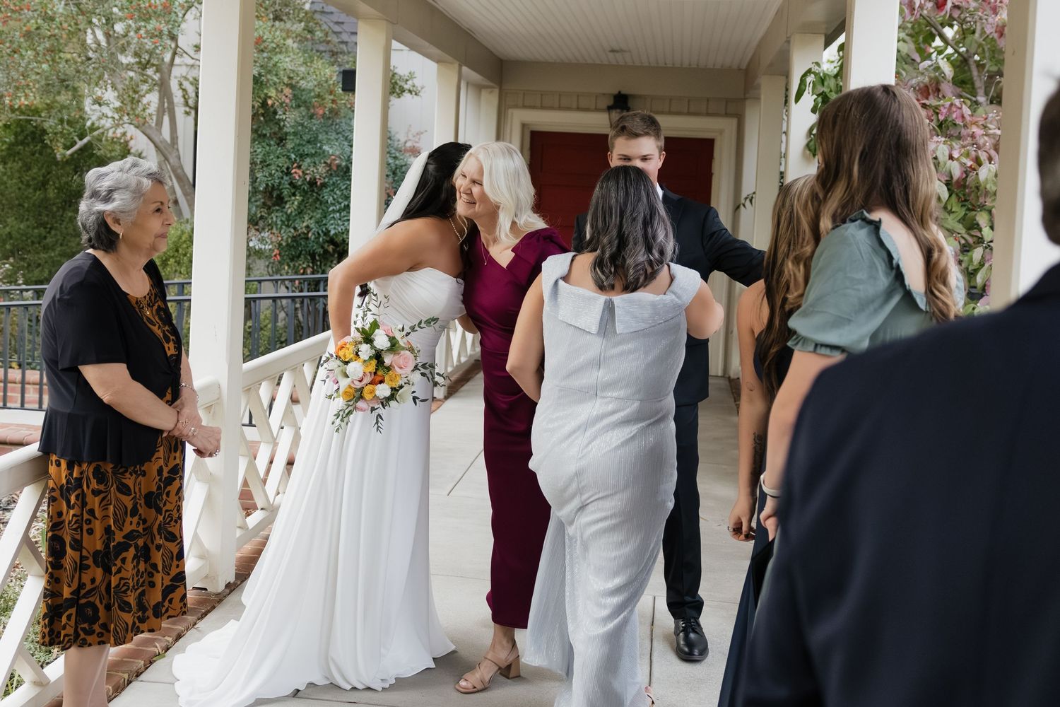 Bride in white gown receiving warm congratulations from guests on a porch after wedding ceremony.