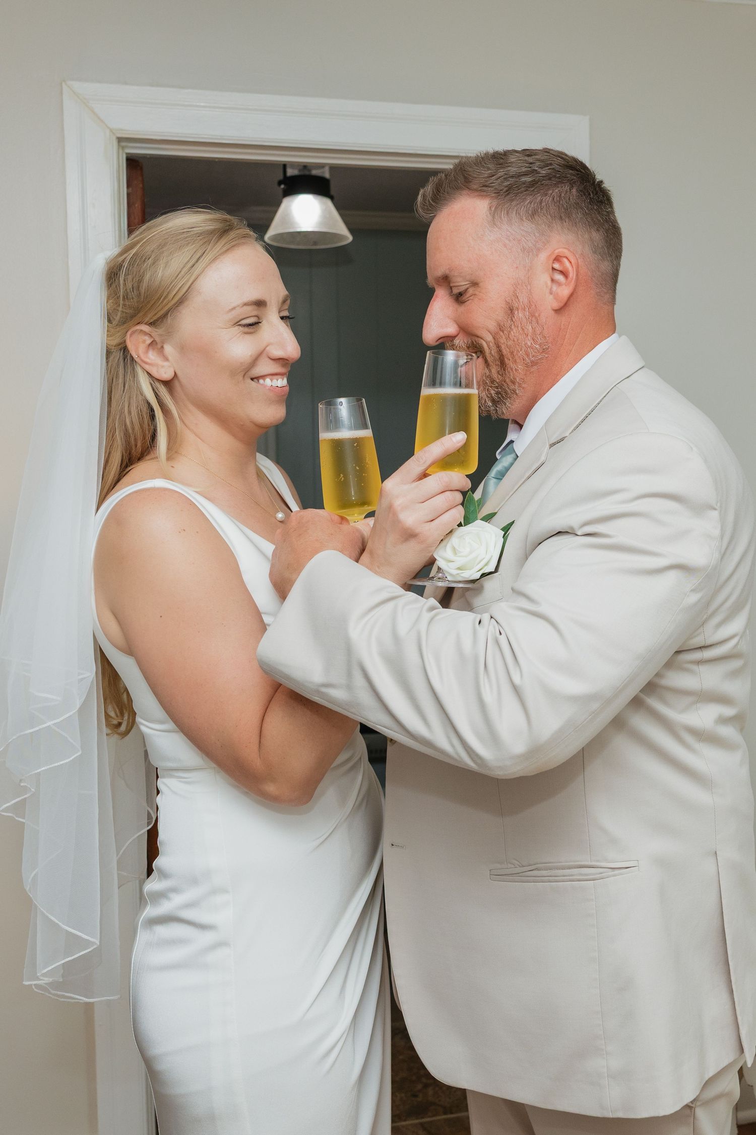 Bride and groom in beige suit share a champagne toast, smiling warmly at each other indoors.