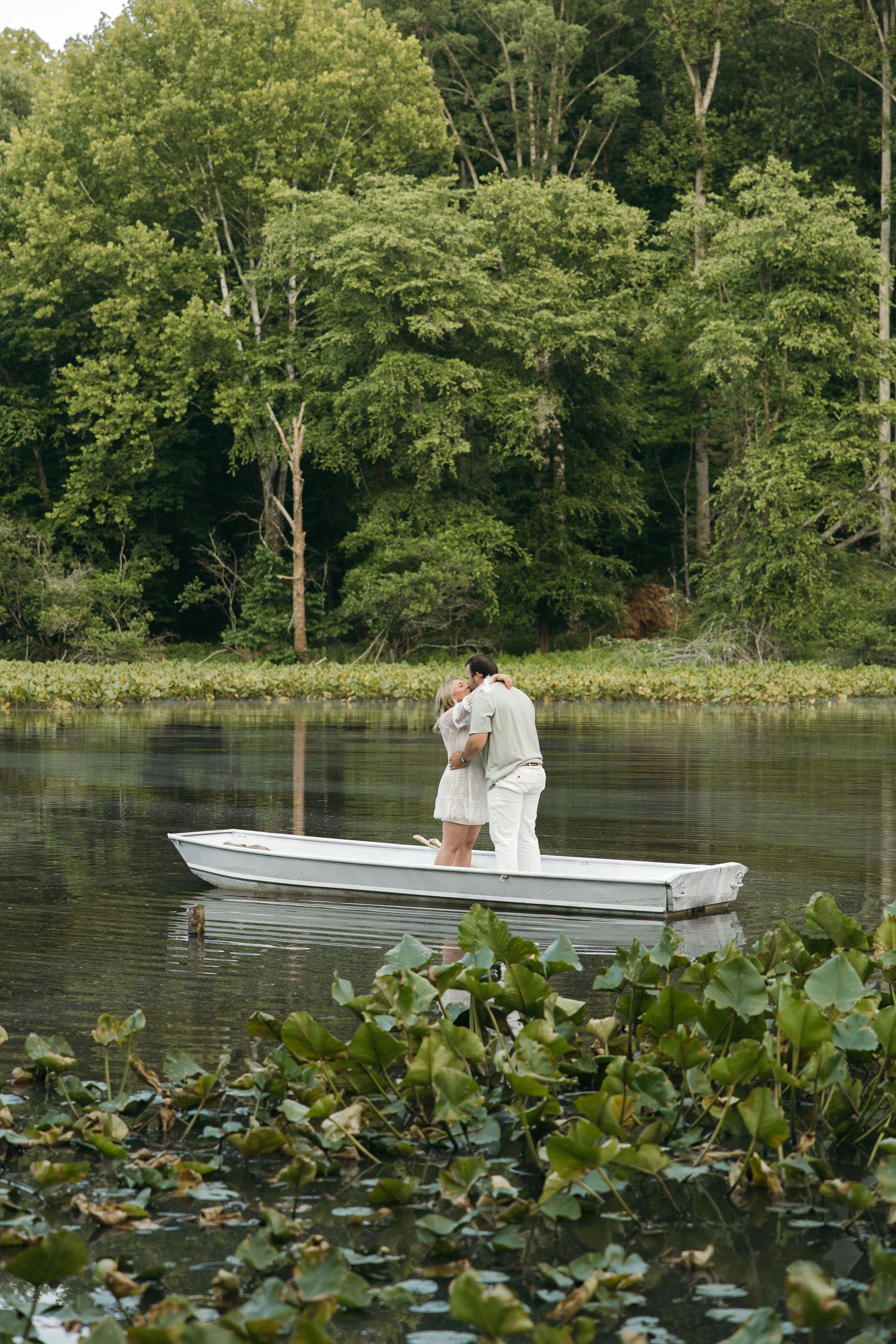 Romantic couple embracing while standing in a white rowboat on a calm river with lily pads.