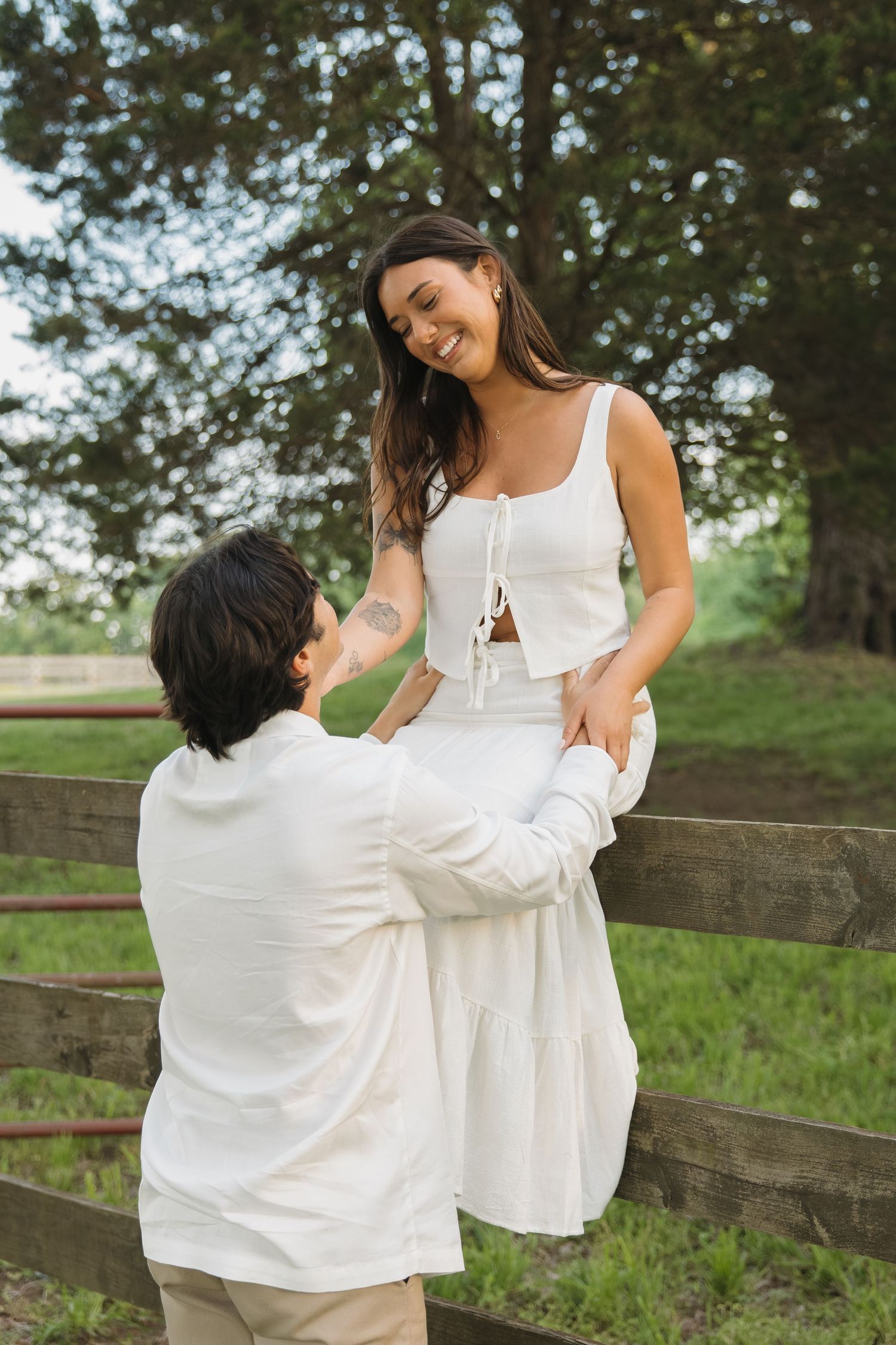 Smiling woman sitting on a wooden fence while her partner holds her hands in a green park.