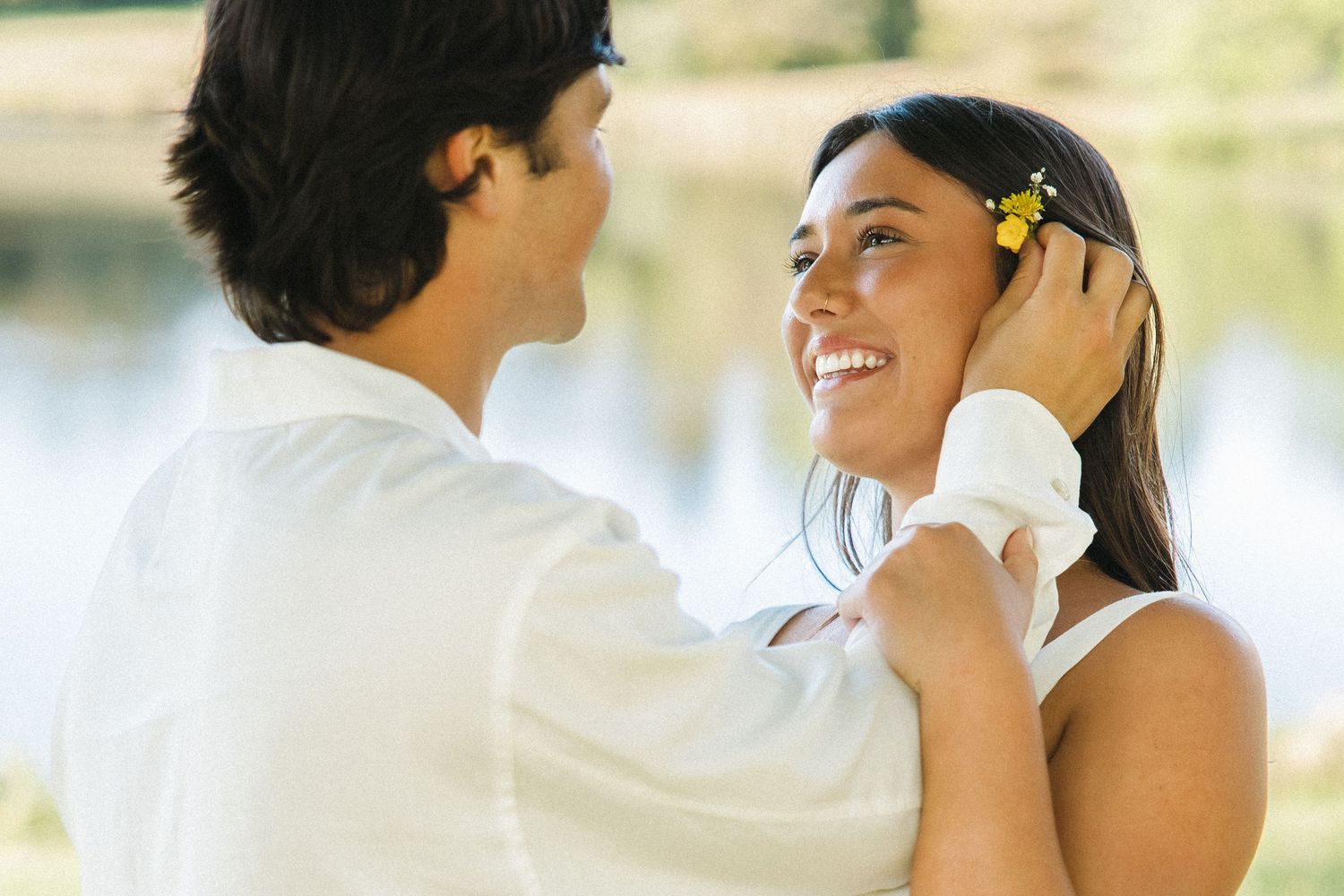 Smiling woman with yellow flower in hair gazes at partner while he gently cups her face near a lake.