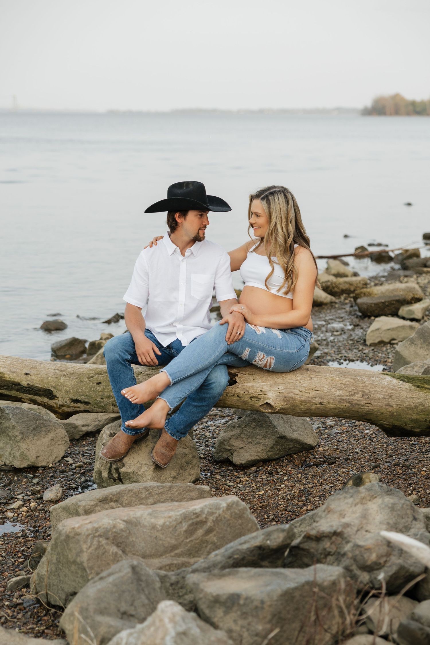 Smiling couple sitting on driftwood by rocky shoreline, man in black cowboy hat gazing at pregnant partner.