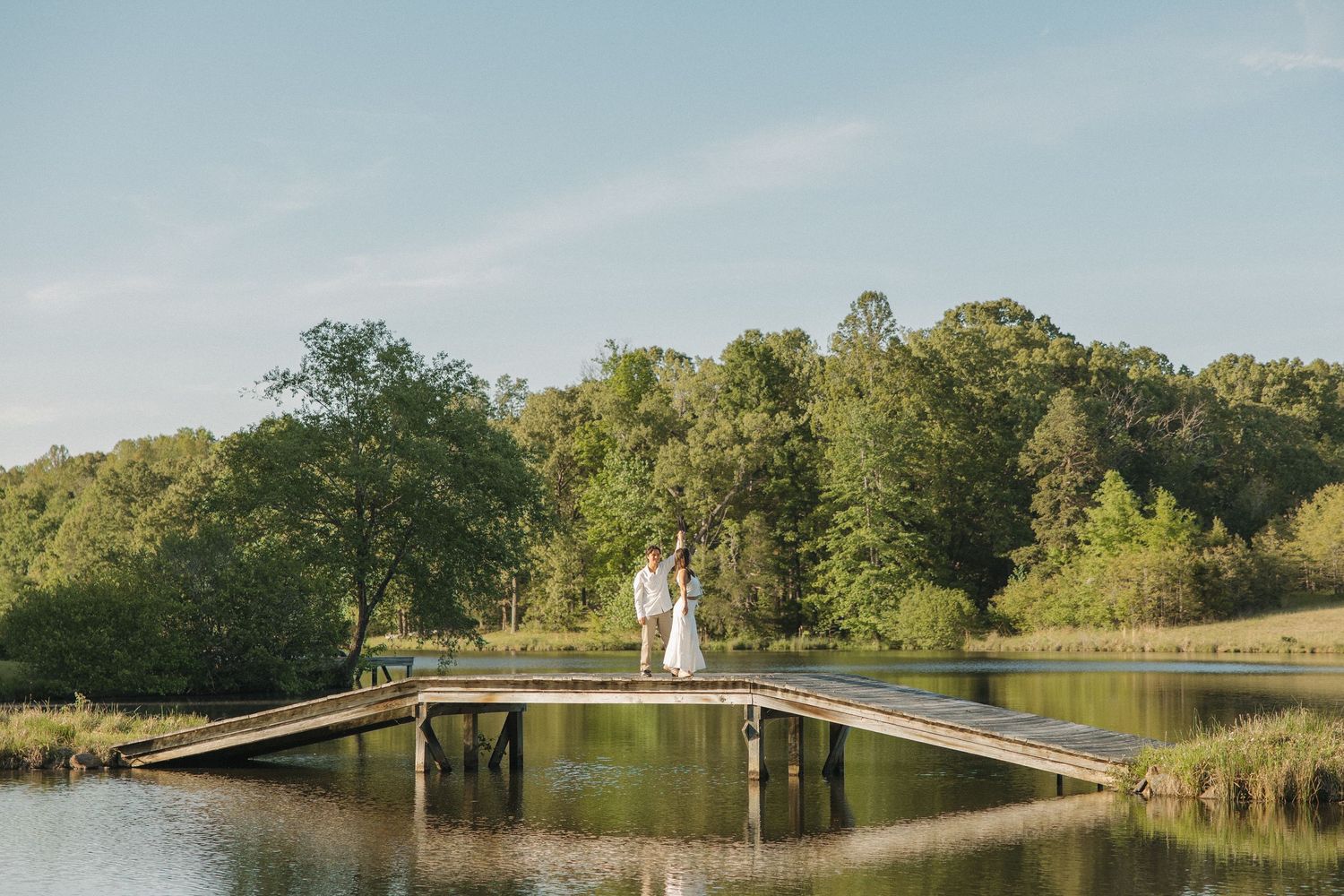 Bride and groom standing together on a wooden pier over a serene lake with green forest backdrop.